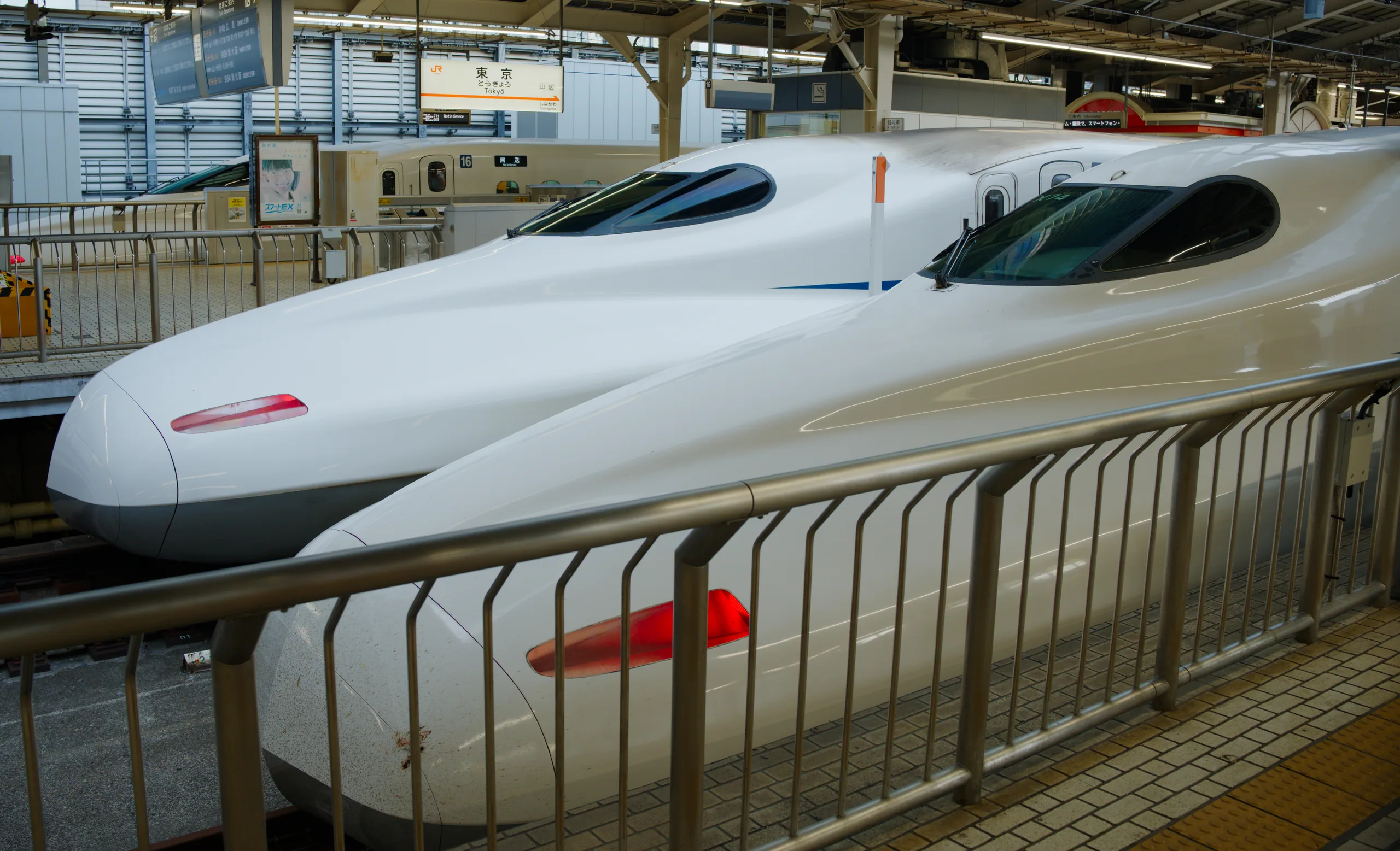Multiple white bullet trains line a train station platform with metal railings and overhead structural beams.