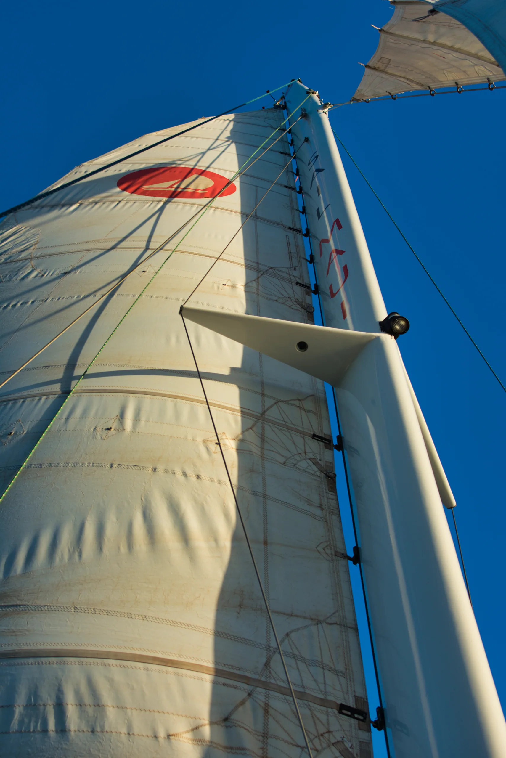 A yacht's mainsail seen from the deck looking up the mast against a deep blue sky.