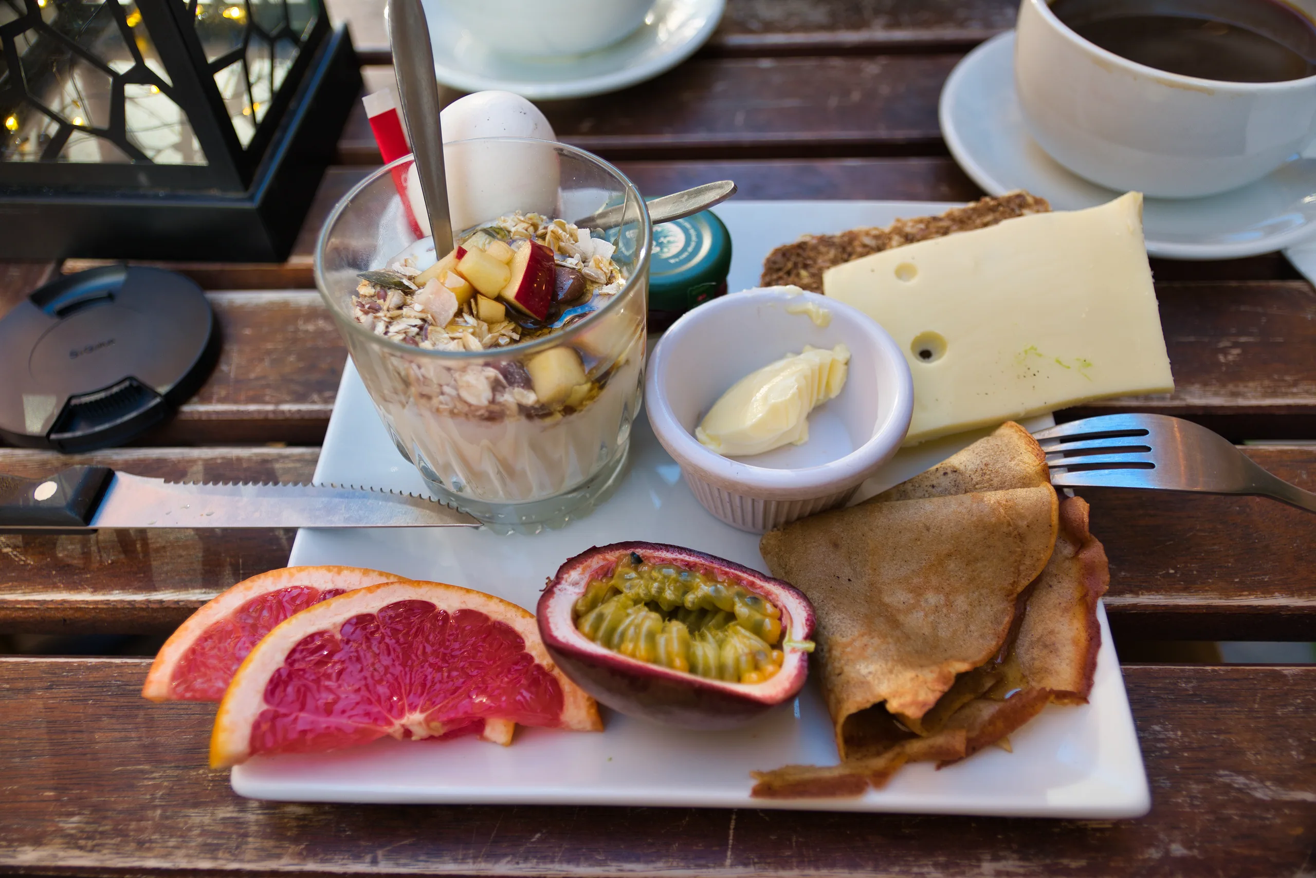 A cafe breakfast on a wooden table — muesli in a glass, cheese and cold cuts on a white plate, grapefruit and passion fruit, and a cup of coffee.