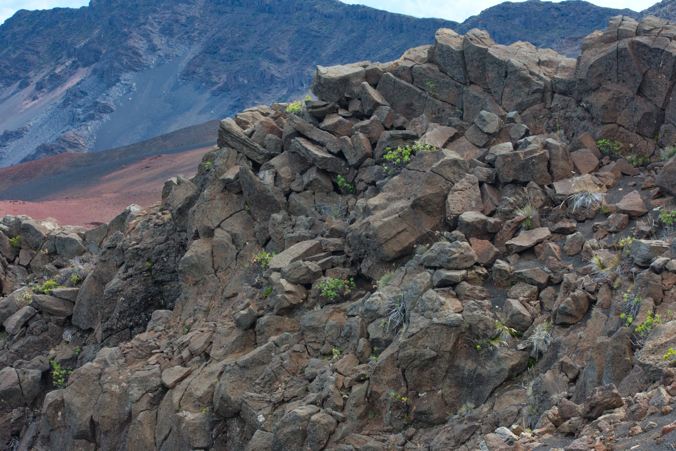 Jagged blocks of dark lava in the foreground with the eroded red and grey slopes of a crater rising behind.