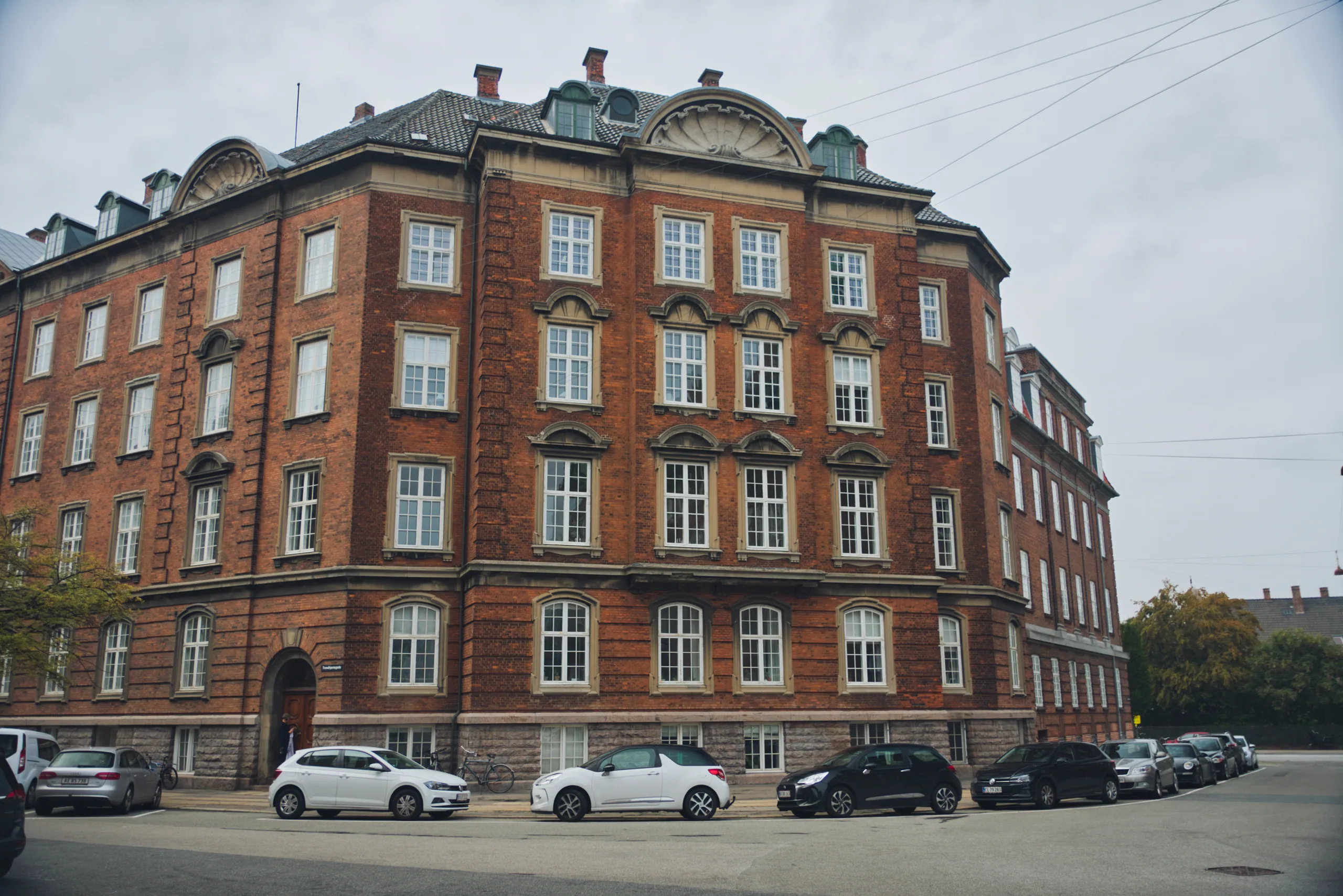 A five-storey red-brick apartment building on a corner, its windows picked out in white stone, cars parked along the street below.