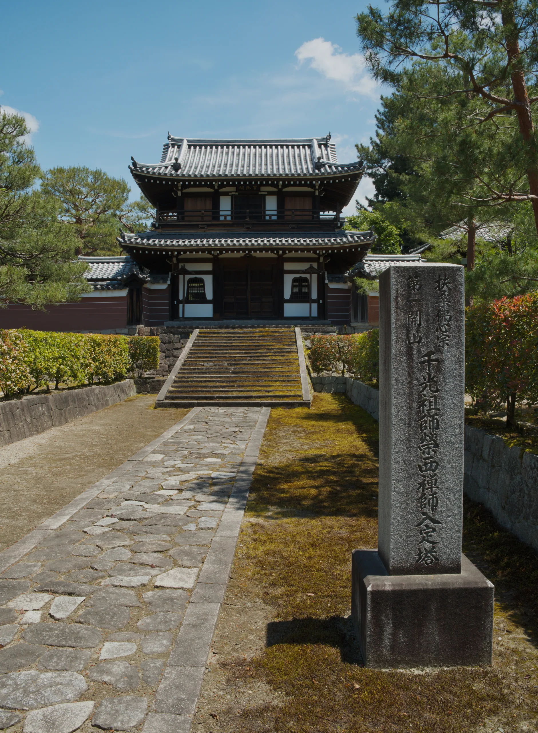 A stone pathway and carved pillar lead toward a traditional East Asian pagoda with tiered roofs set among moss and trees.