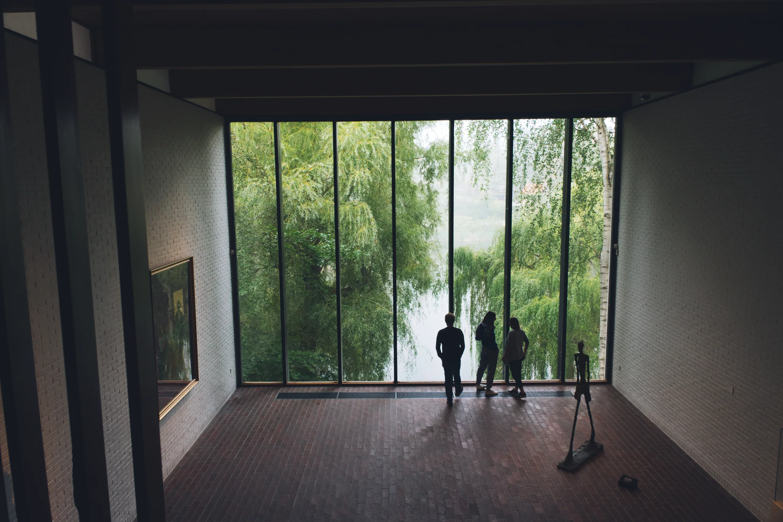 A darkened gallery looking out through a full-height window onto willows and water, thin standing figure sculptures silhouetted against the light.