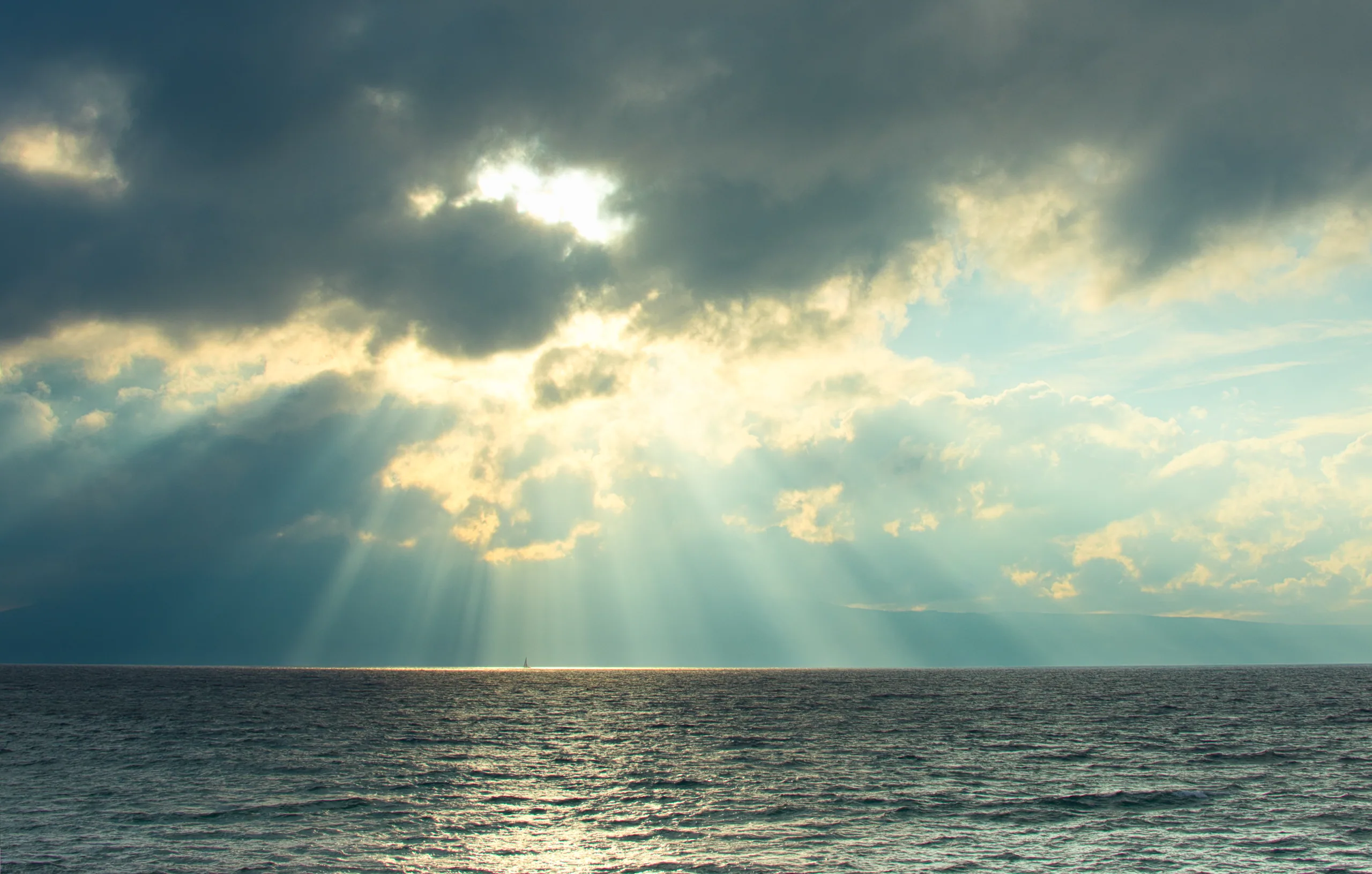 Shafts of sunlight breaking through heavy cloud over a dark open ocean.