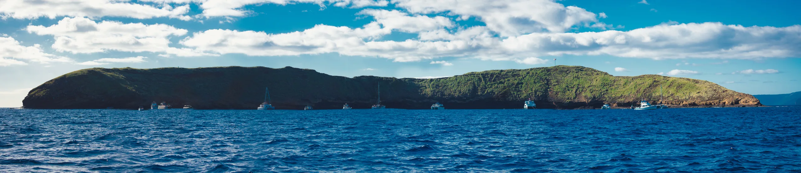 The long low crescent of Molokini on the horizon under scattered cloud, boats moored along its inner shore, seen across deep blue water.