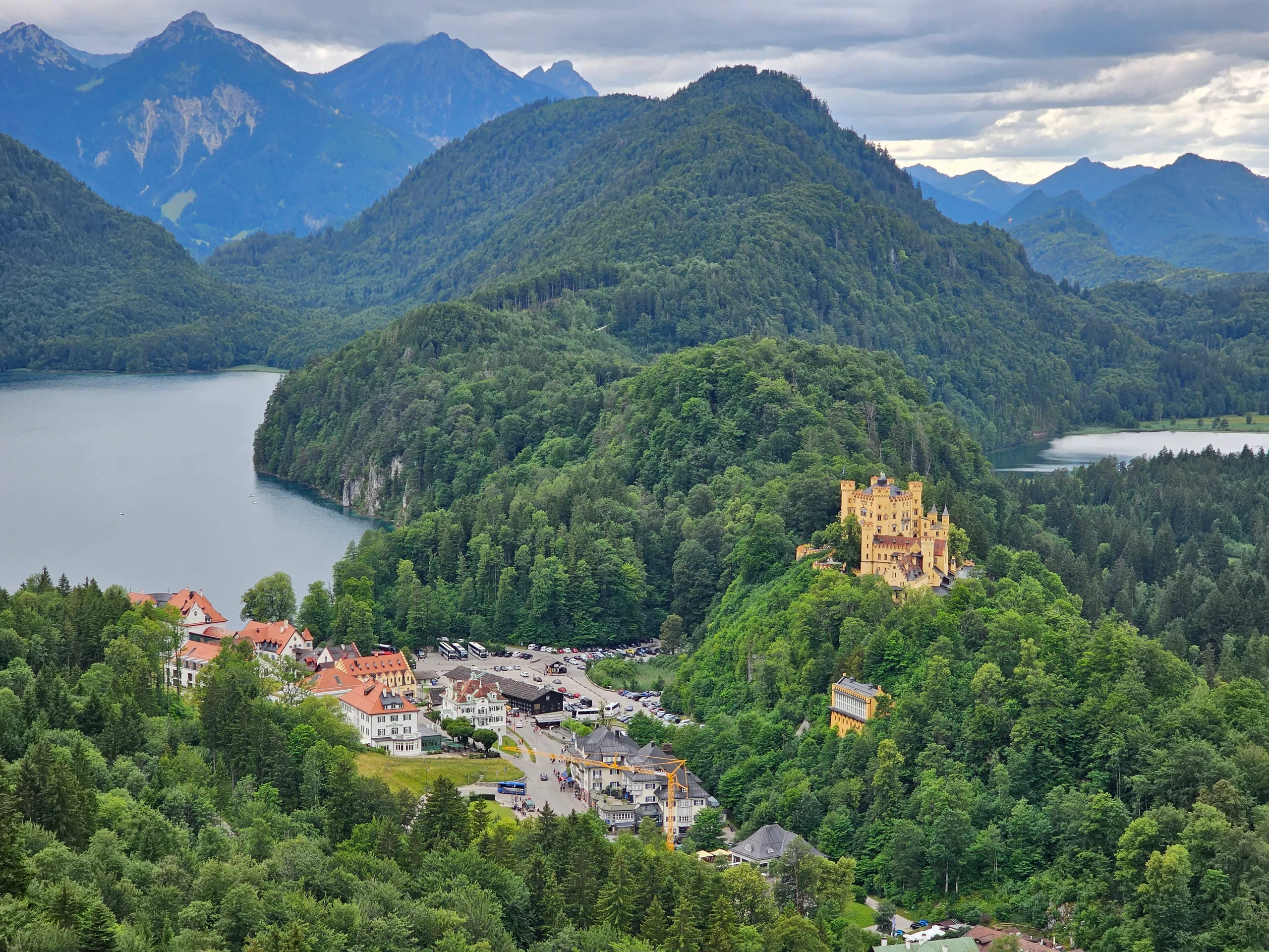 A view over Hohenschwangau village and its lake to forested mountains, the yellow castle among the trees.