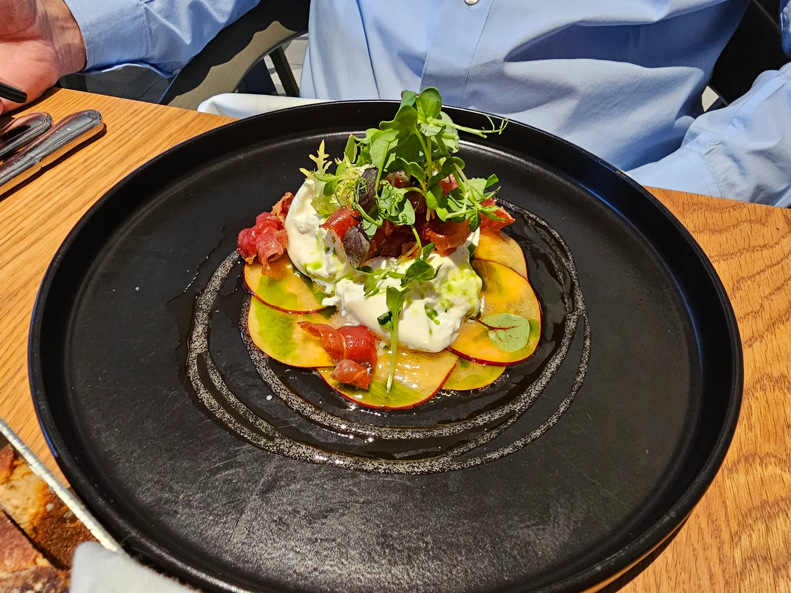 A dish of thinly sliced beetroot and tomato under microgreens and a white sauce, on a black plate.