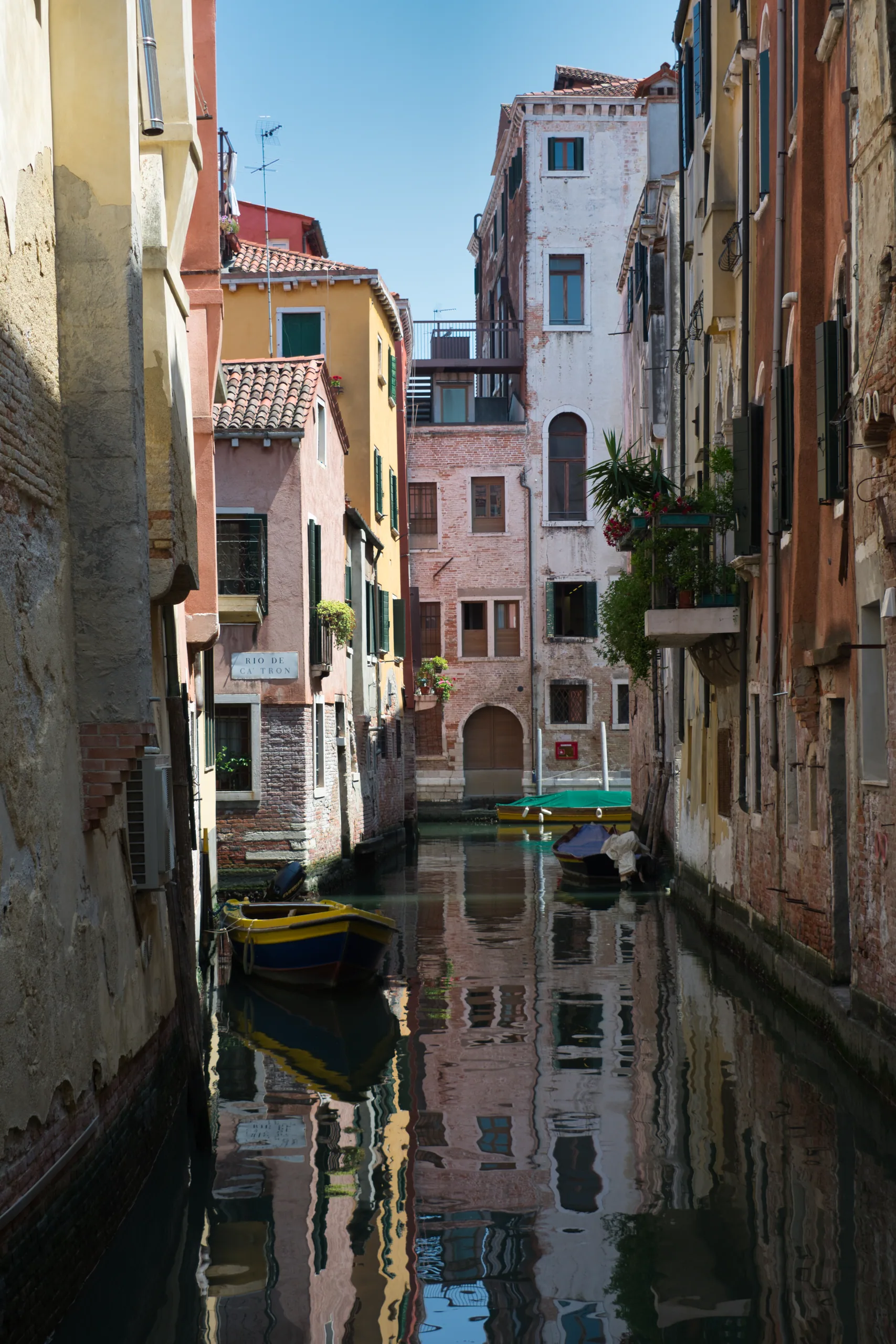 A narrow Venetian canal between tall stuccoed houses, small boats moored along it and the buildings mirrored in still green water.