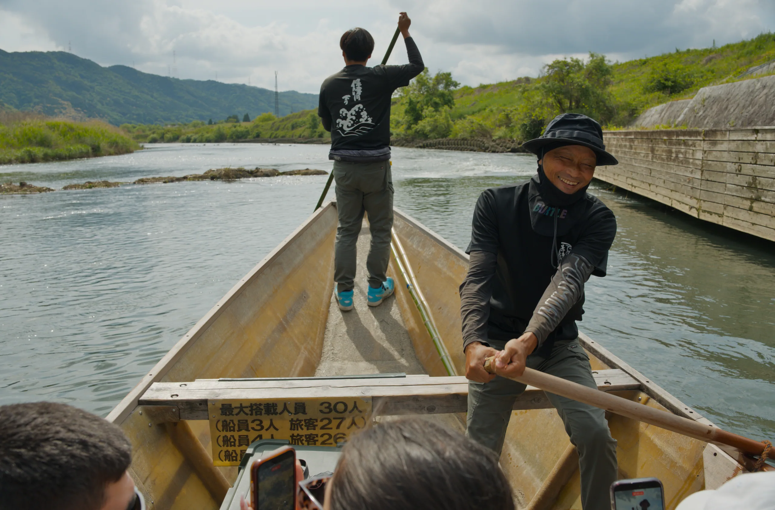 A person stands with raised arms on a boat's bow, river and mountains stretching behind.