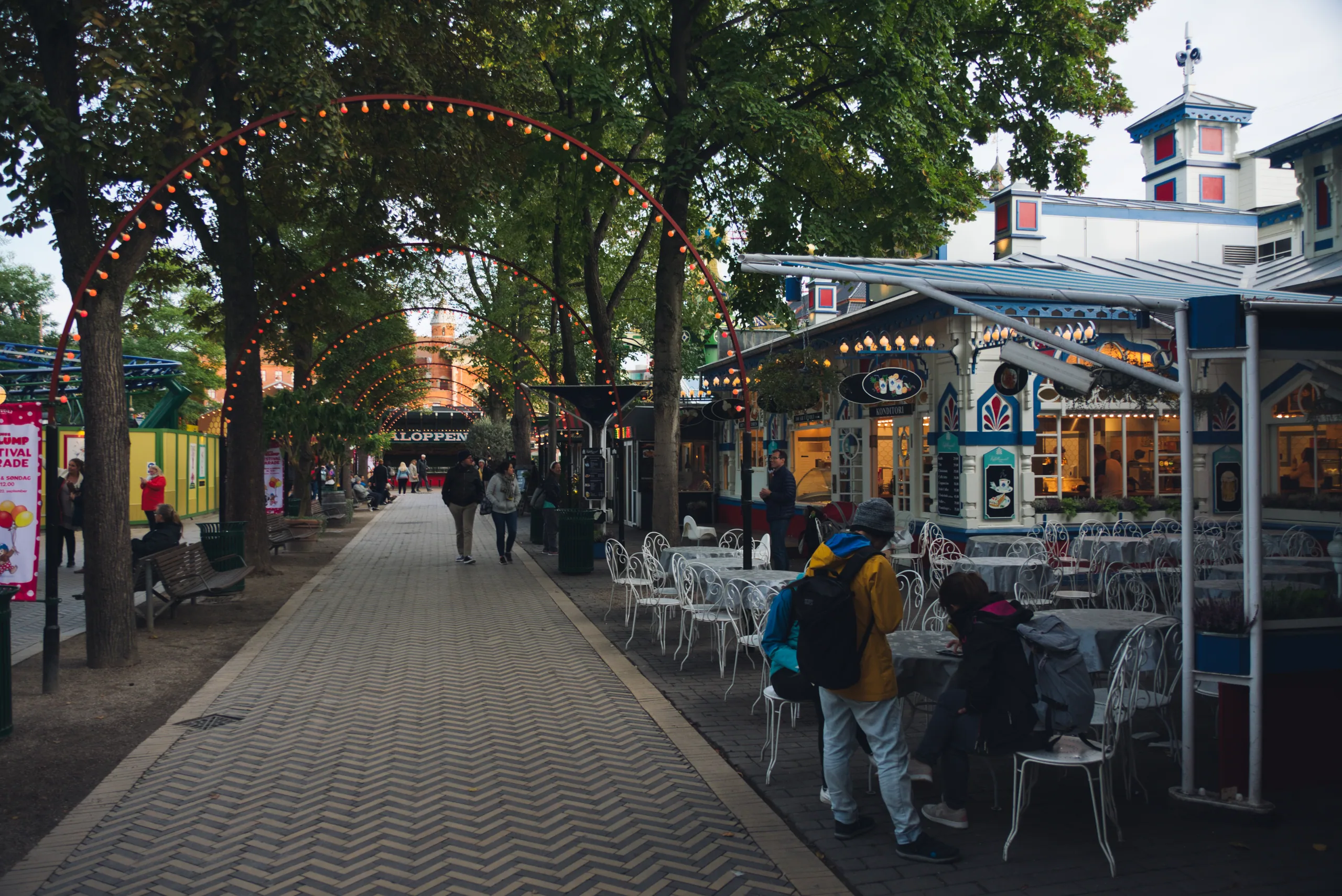 A tree-lined avenue through Tivoli strung with arches of lights, cafe tables and pavilions along one side.