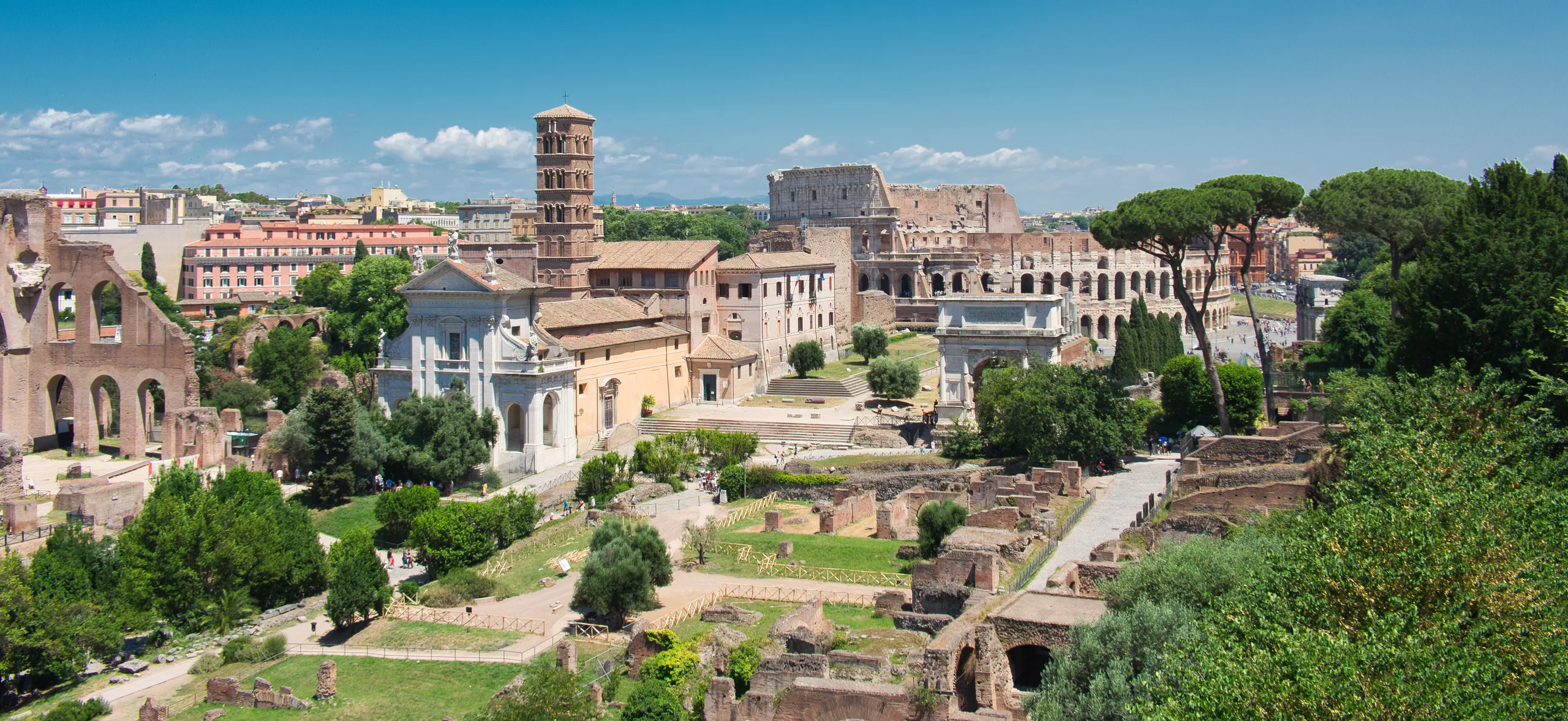 A view over the Roman Forum from above, ruined columns and foundations among trees with the Colosseum beyond.