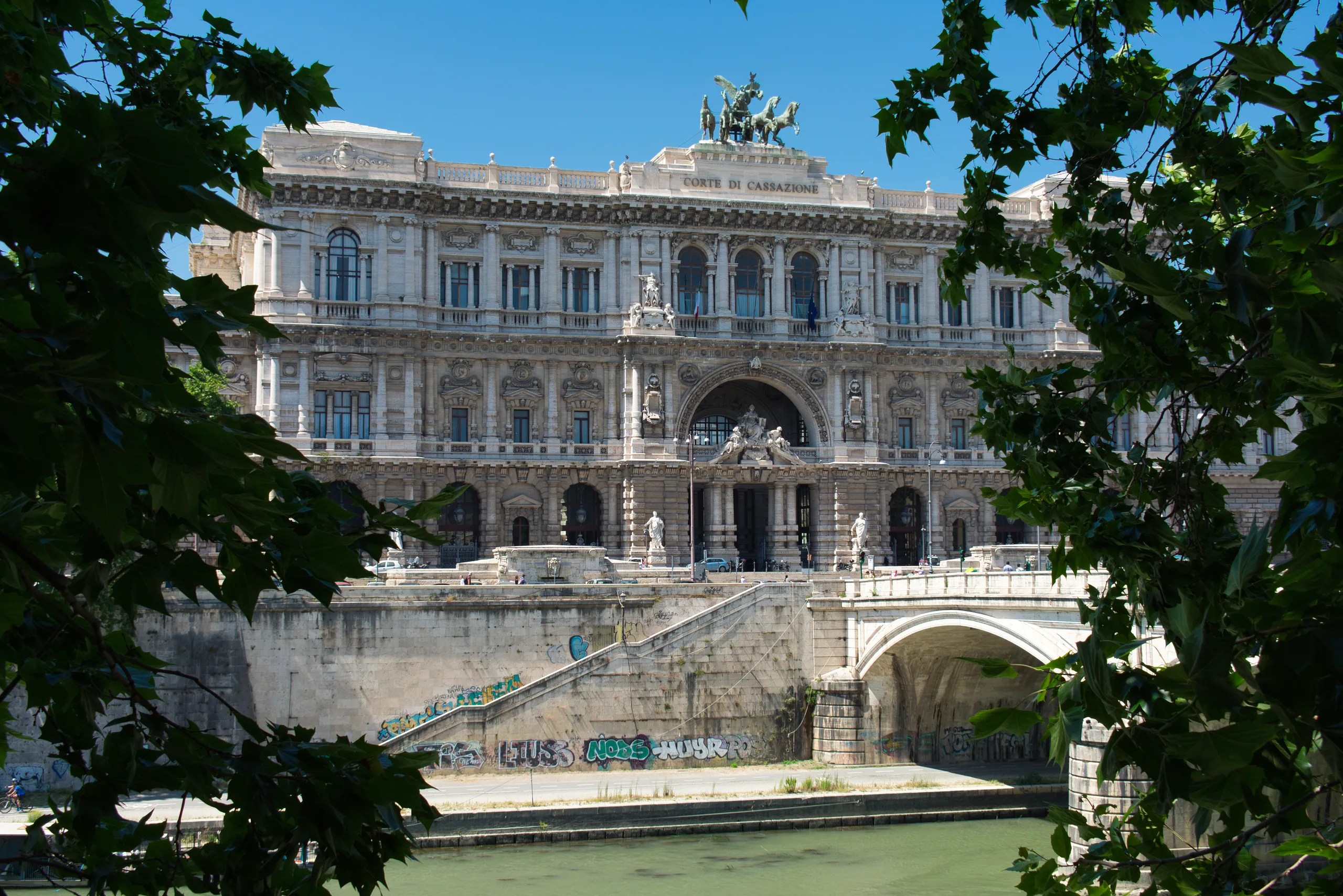 The Palace of Justice in Rome seen through a frame of leaves, its sculpted facade above the river embankment.