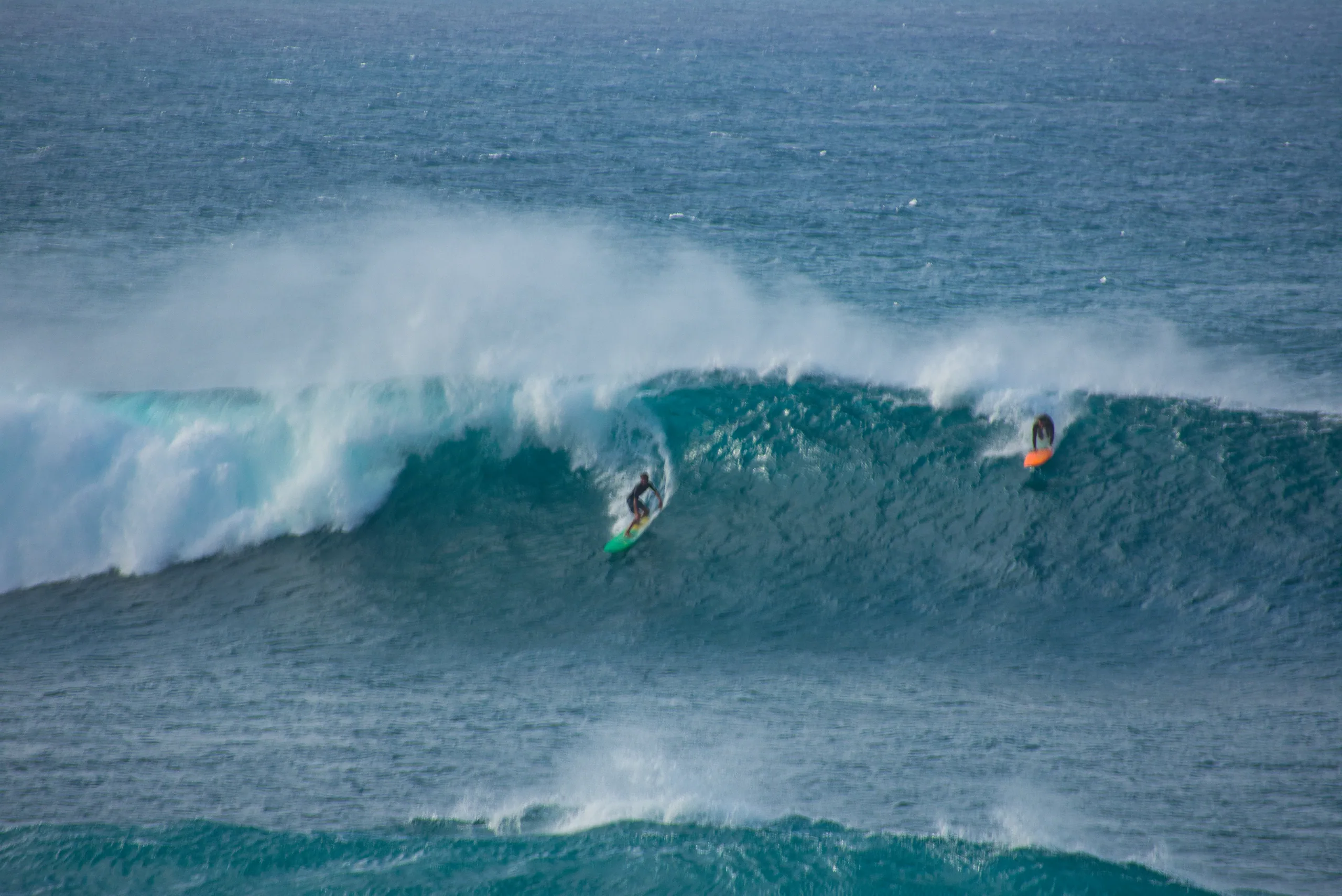 A surfer riding inside the curl of a large breaking wave, another paddling over the shoulder behind.