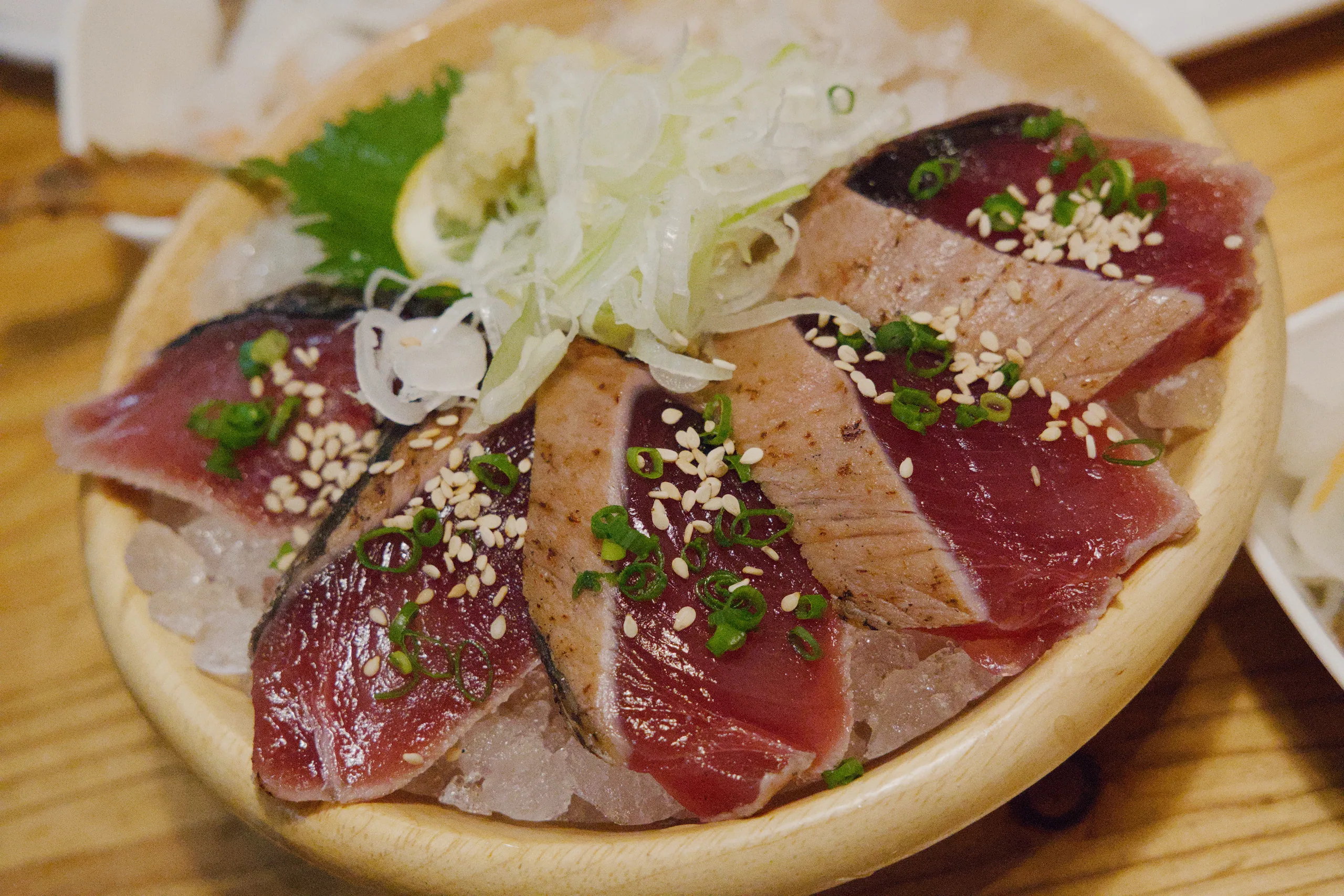 Seared bonito sashimi fanned over crushed ice in a pale wooden bowl, scattered with sesame seeds and sliced spring onion, a nest of shredded leek behind it.