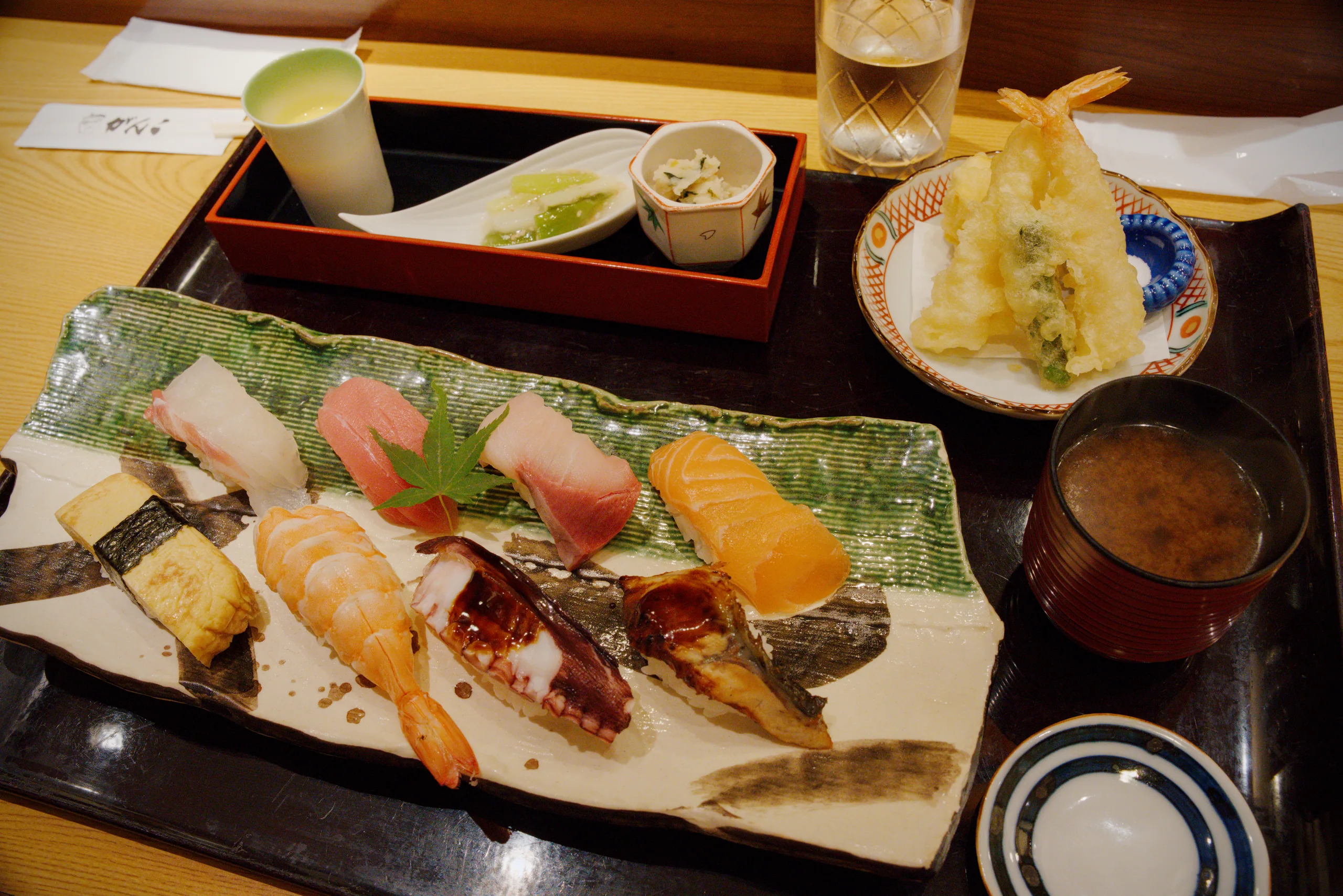 Japanese meal with sushi, grilled items, tempura, rice, and soup in black lacquered compartments.