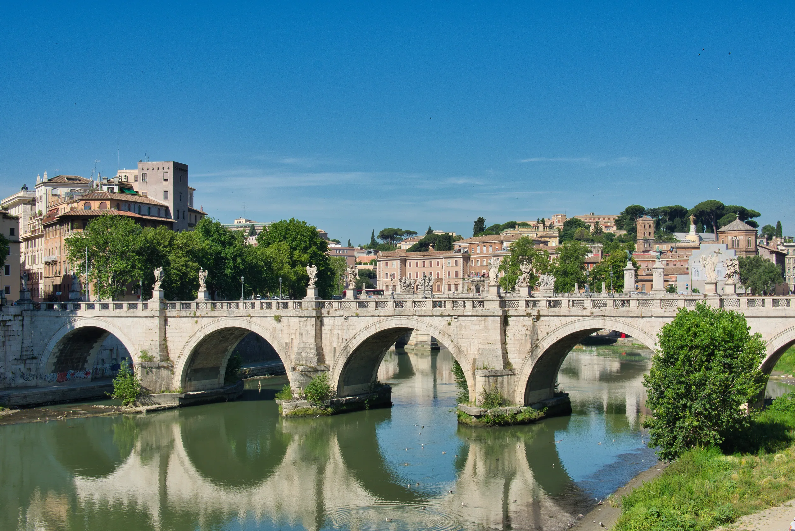 An arched stone bridge over the green Tiber, the rooftops and towers of Rome rising behind it.
