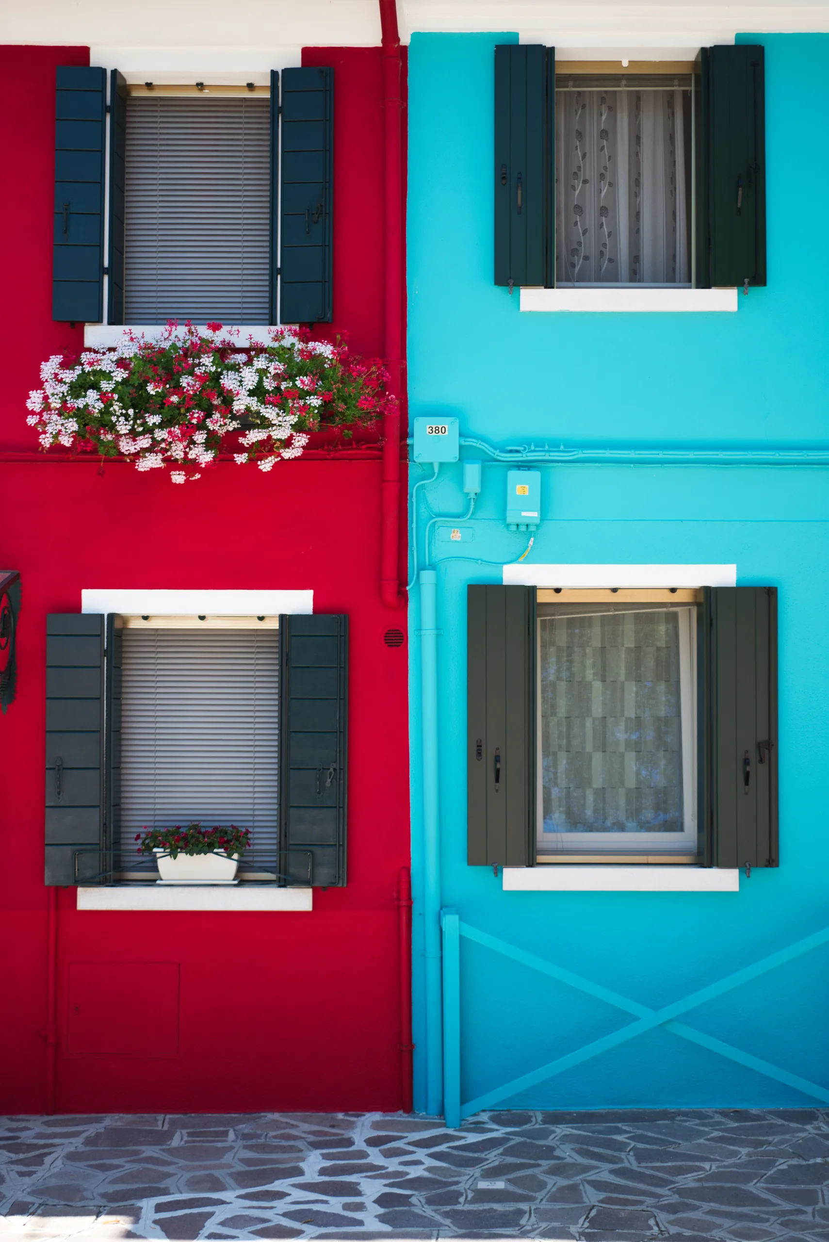 Two adjoining house fronts on Burano, one scarlet and one turquoise, with grey shutters and a window box of pink flowers.