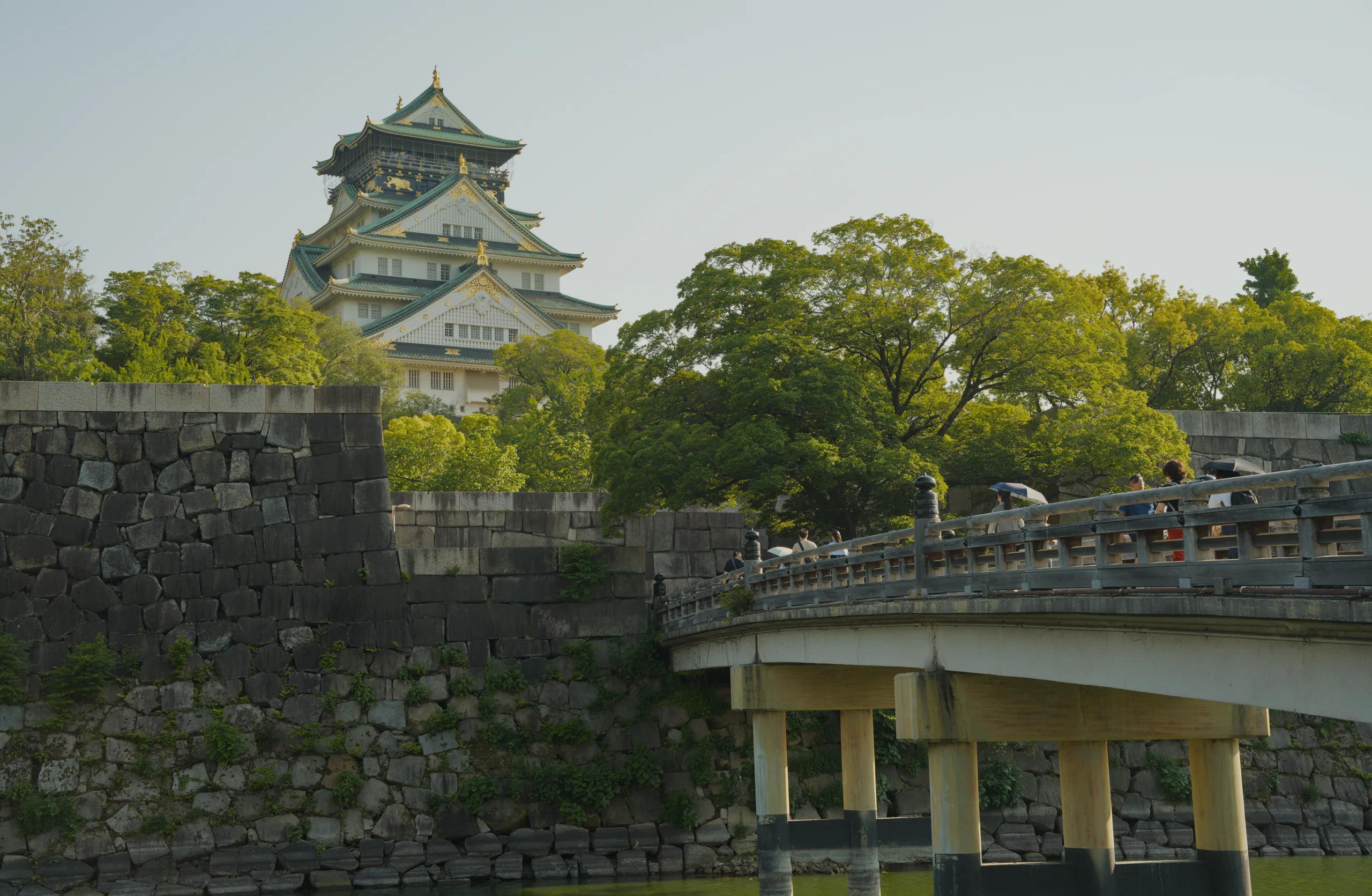 Japanese castle with tiered tower overlooking stone walls, moat, and visitors on a bridge below.