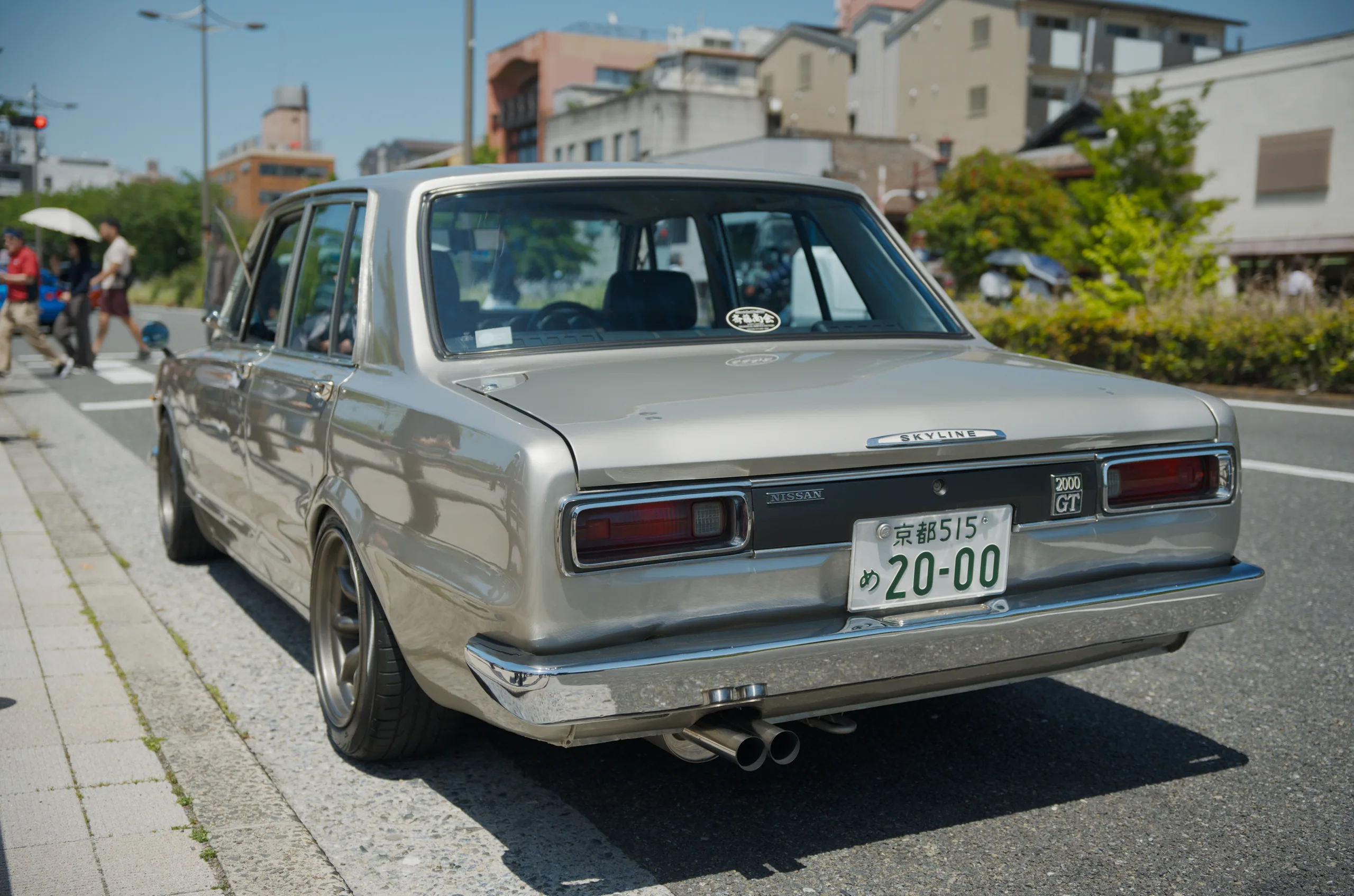 A white vintage sedan with a Japanese license plate sits parked on a street lined with buildings and pedestrians.