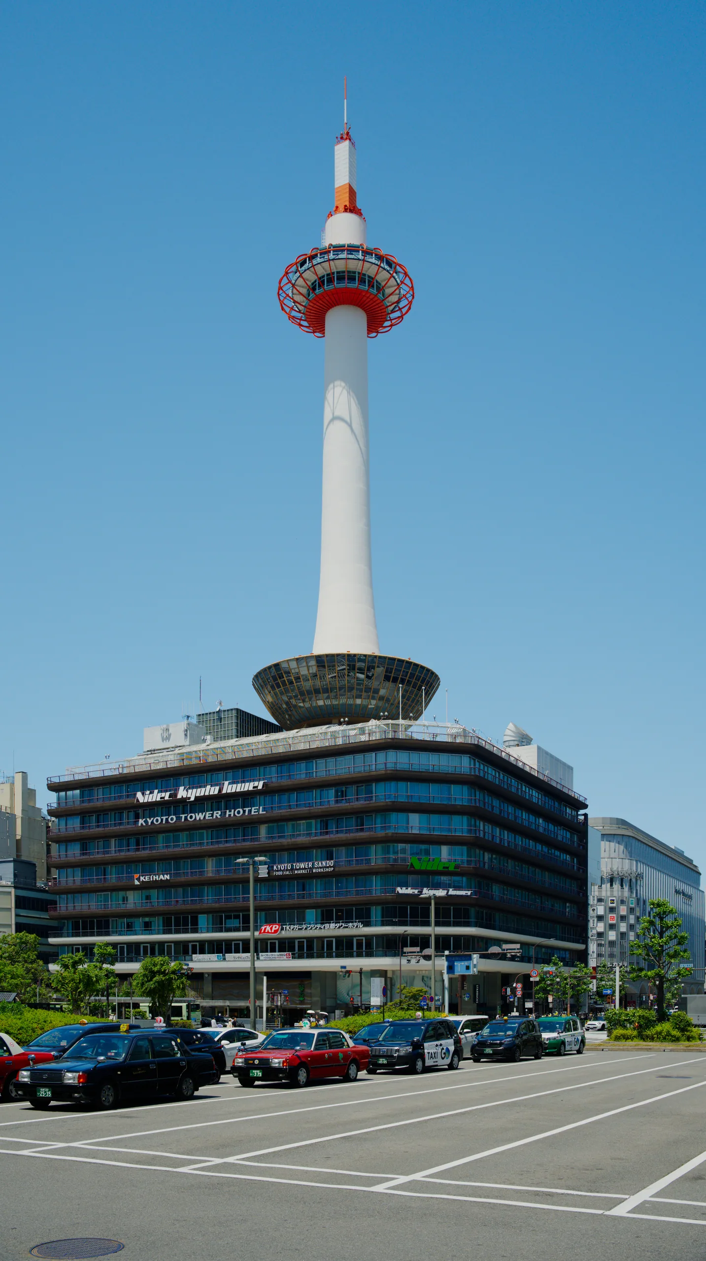 A white tower with a red pagoda-style observation deck rises above a modern building and parking lot.