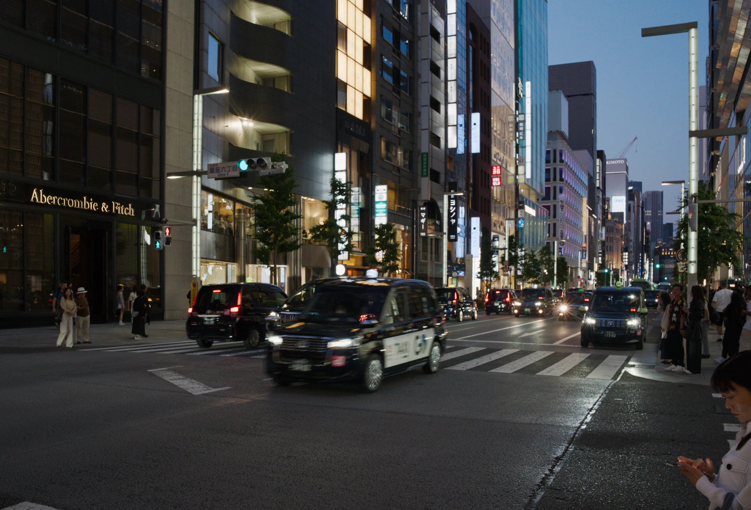 Urban street at night with illuminated storefronts, vehicles, and tall buildings.
