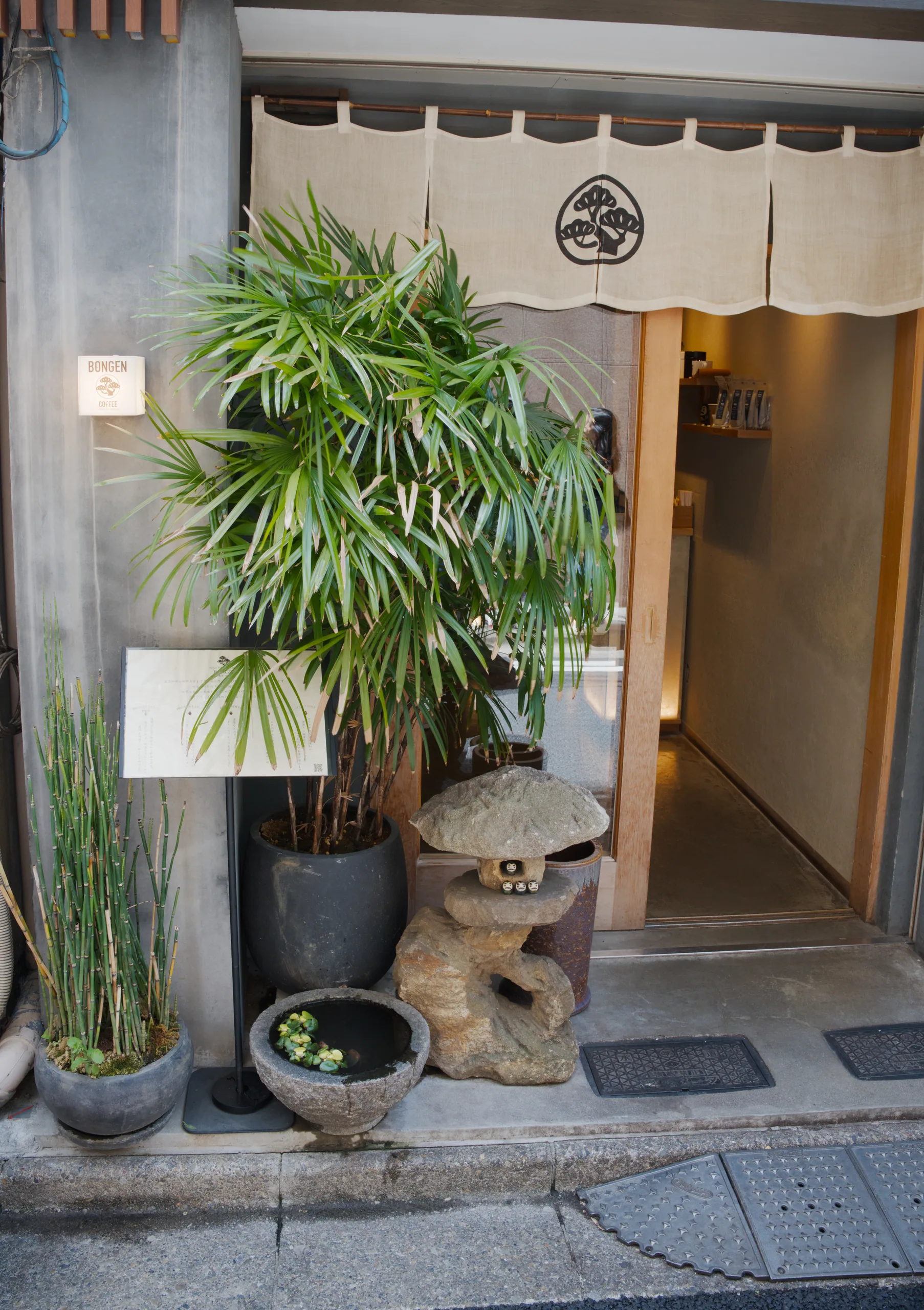 A shop entrance with a large potted palm plant, ceramic ornaments, and a fabric noren curtain.