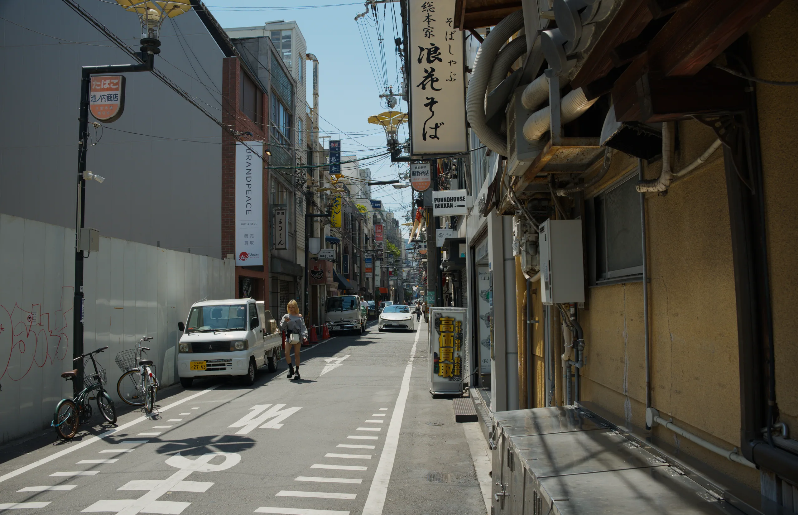 A narrow Japanese street with traditional low buildings, overhead utility wires, and parked vehicles.