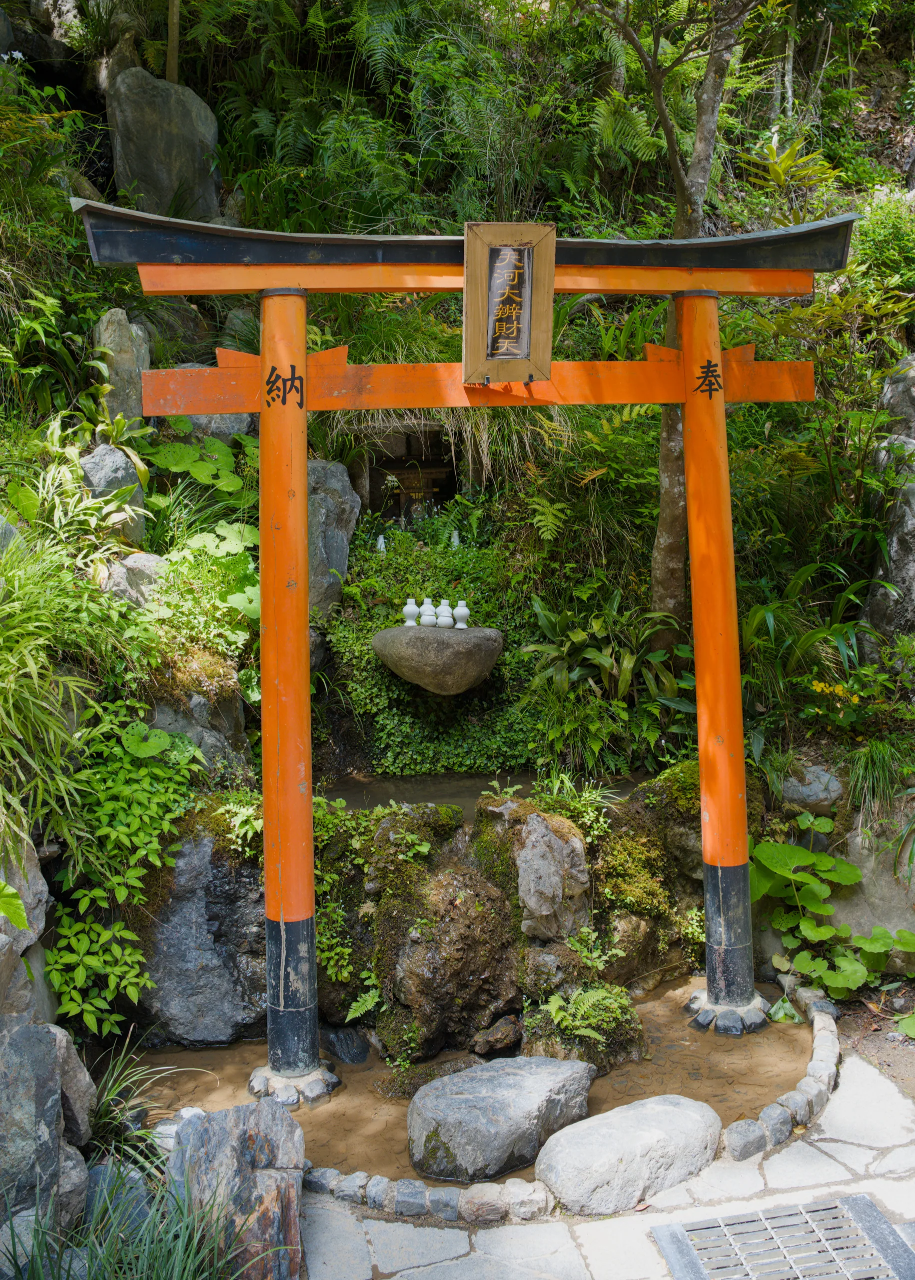 An orange torii gate marks an entrance in a Japanese garden, with stone lanterns and rocks amid dense vegetation.