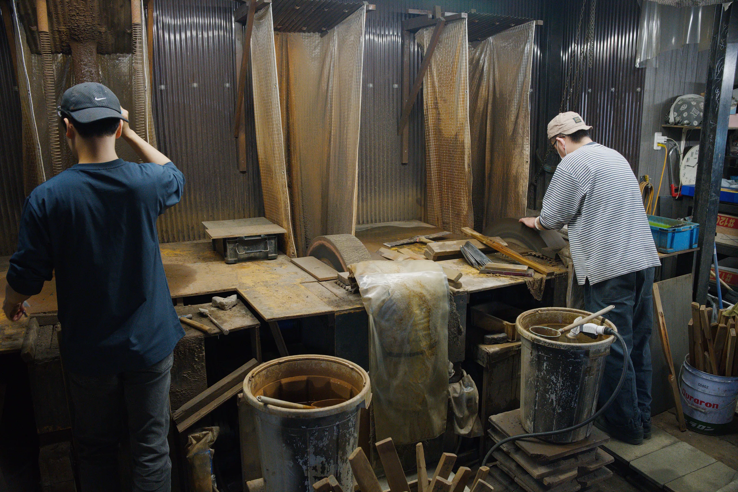Two people work in a workshop with stacked wood and large metal panels, with buckets and hand tools visible.