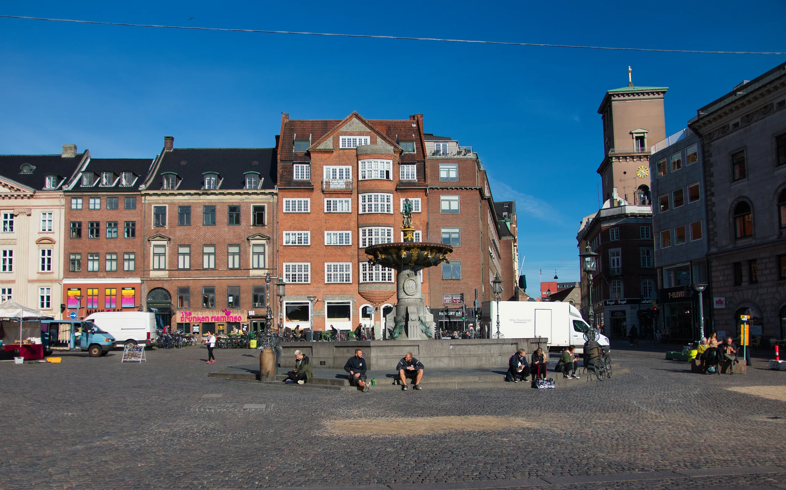 A cobbled Copenhagen square with a fountain, market stalls and cafe awnings along a terrace of tall brick and stucco buildings.