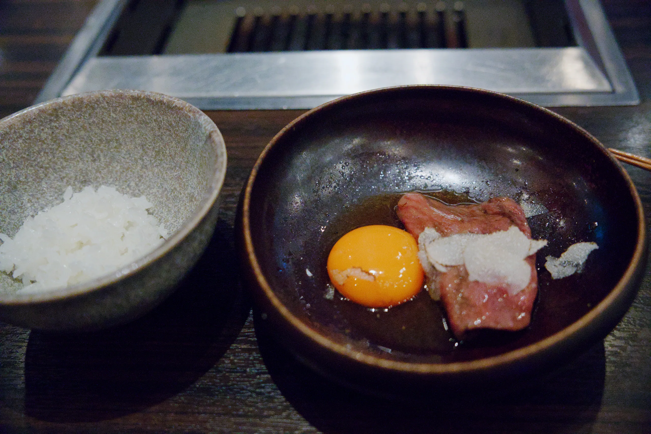 A dark bowl holds a cooked egg and meat on a wooden dining table surrounded by salt, condiments, and dishware.