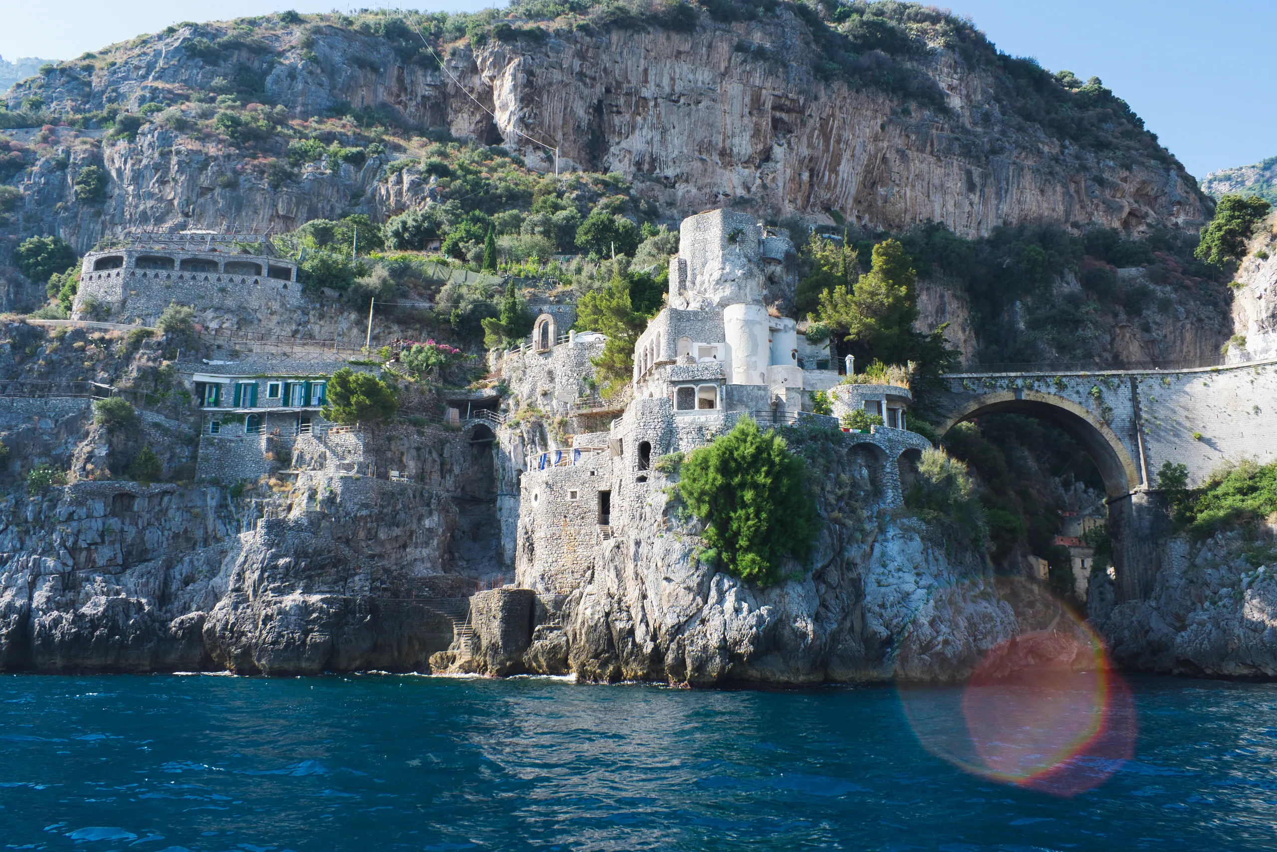 A cliff-side building and stone arch built into the rock above the sea, a lens flare across the frame.
