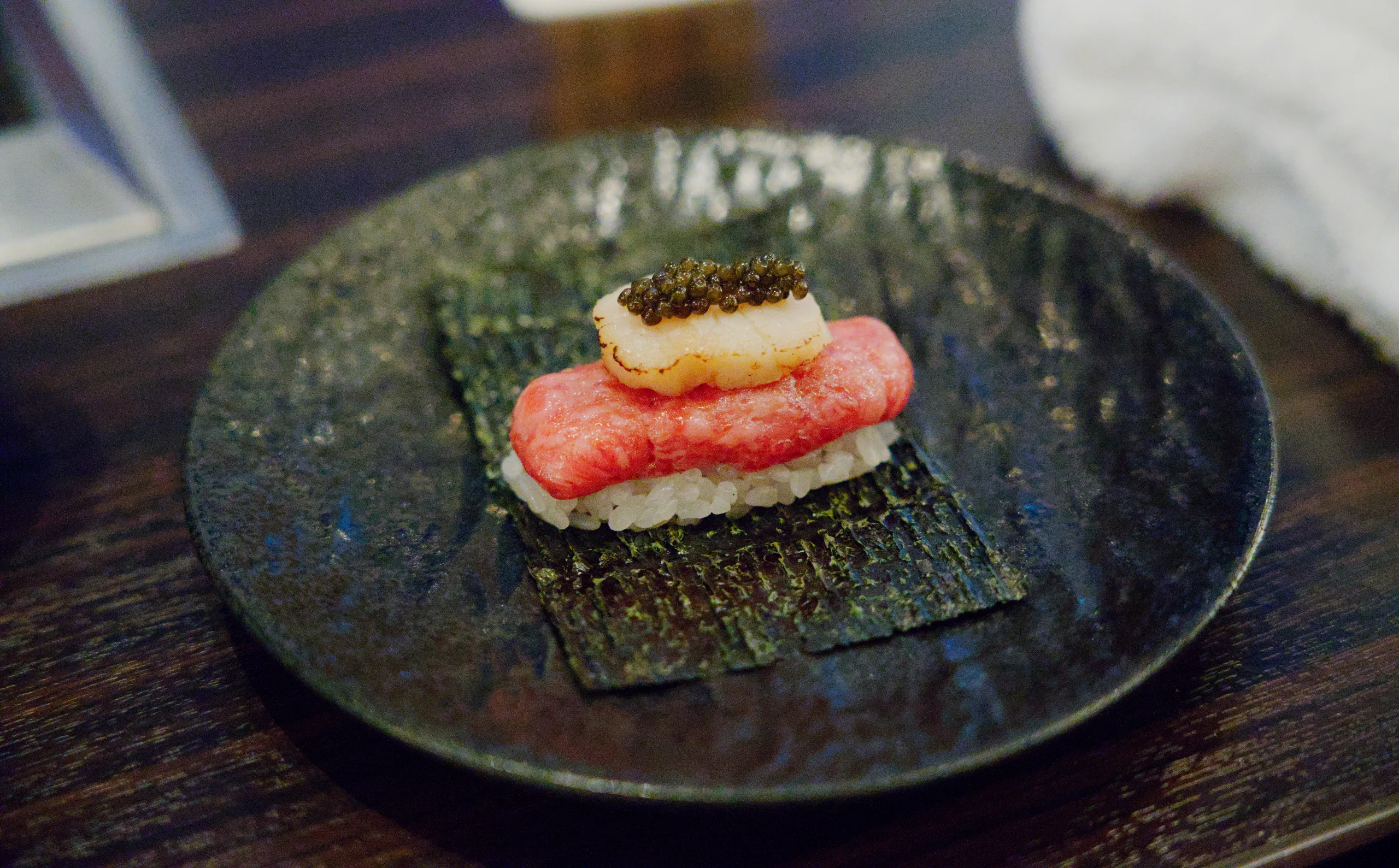 Raw fish, thinly sliced and topped with what appears to be roe and a dark garnish, sits on a textured dark plate.