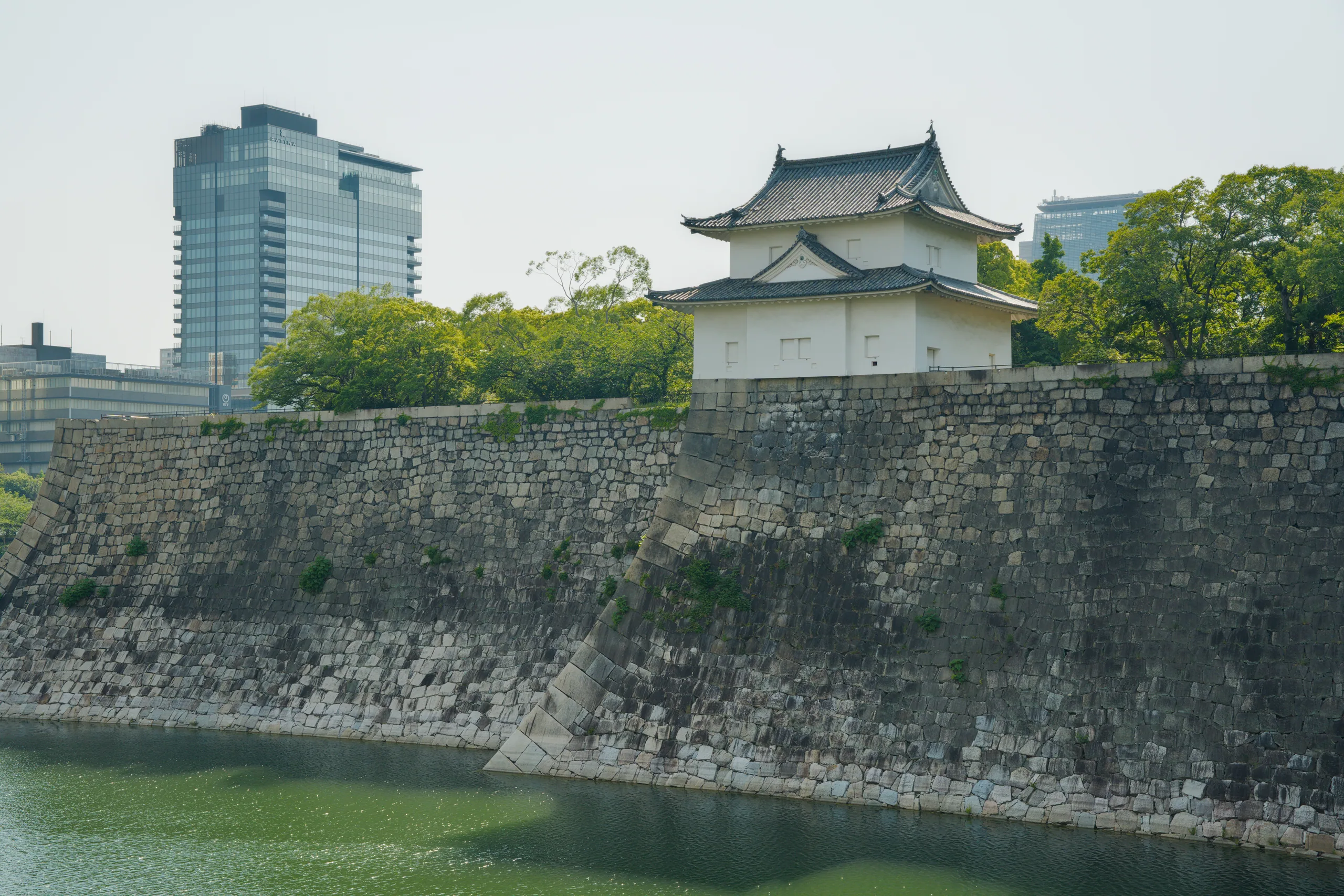 Stone fortification walls angle steeply down to a wide moat with Japanese castle structures visible across the water.
