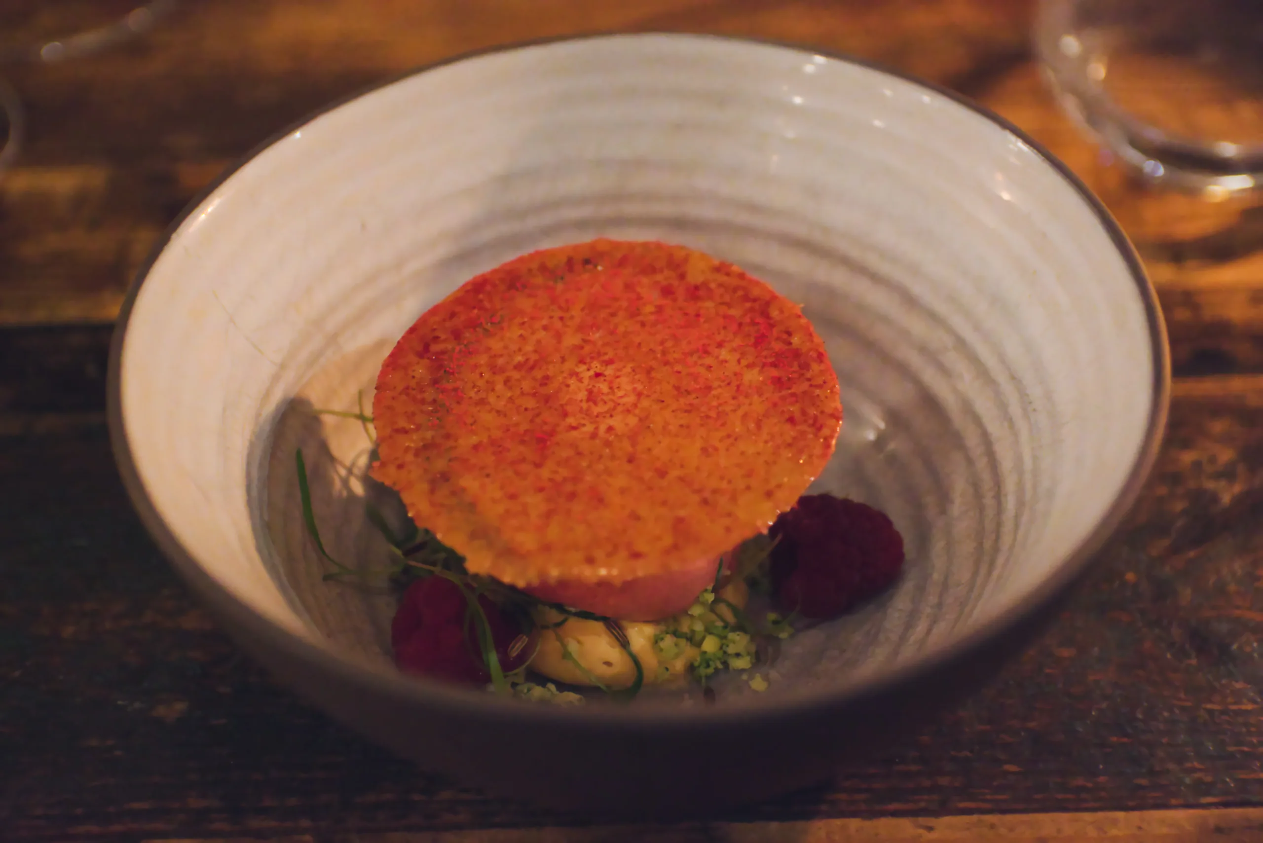 A round tart with a torched orange top sitting in a ridged ceramic bowl, garnished with beetroot and peas.