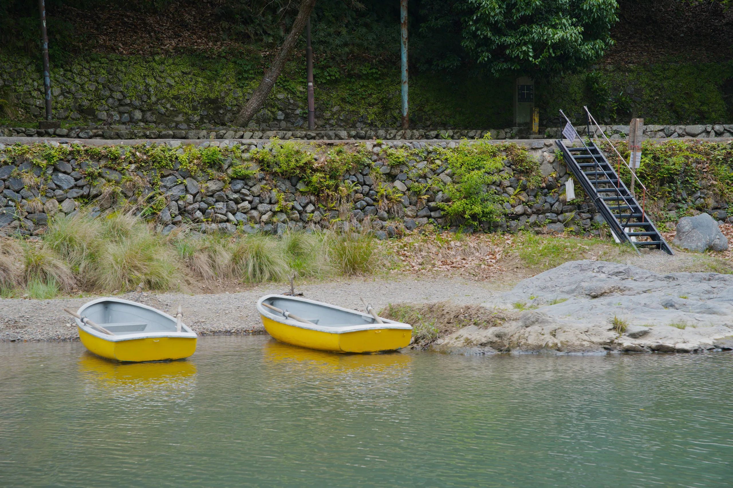 Two yellow boats rest on a pebbled beach below grassy vegetation and a stone wall.