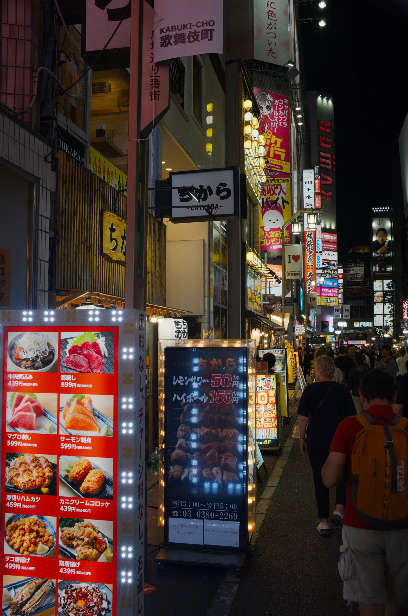 Narrow Japanese alley at night lined with restaurant signage displaying photographs of noodle dishes and small lantern lights.