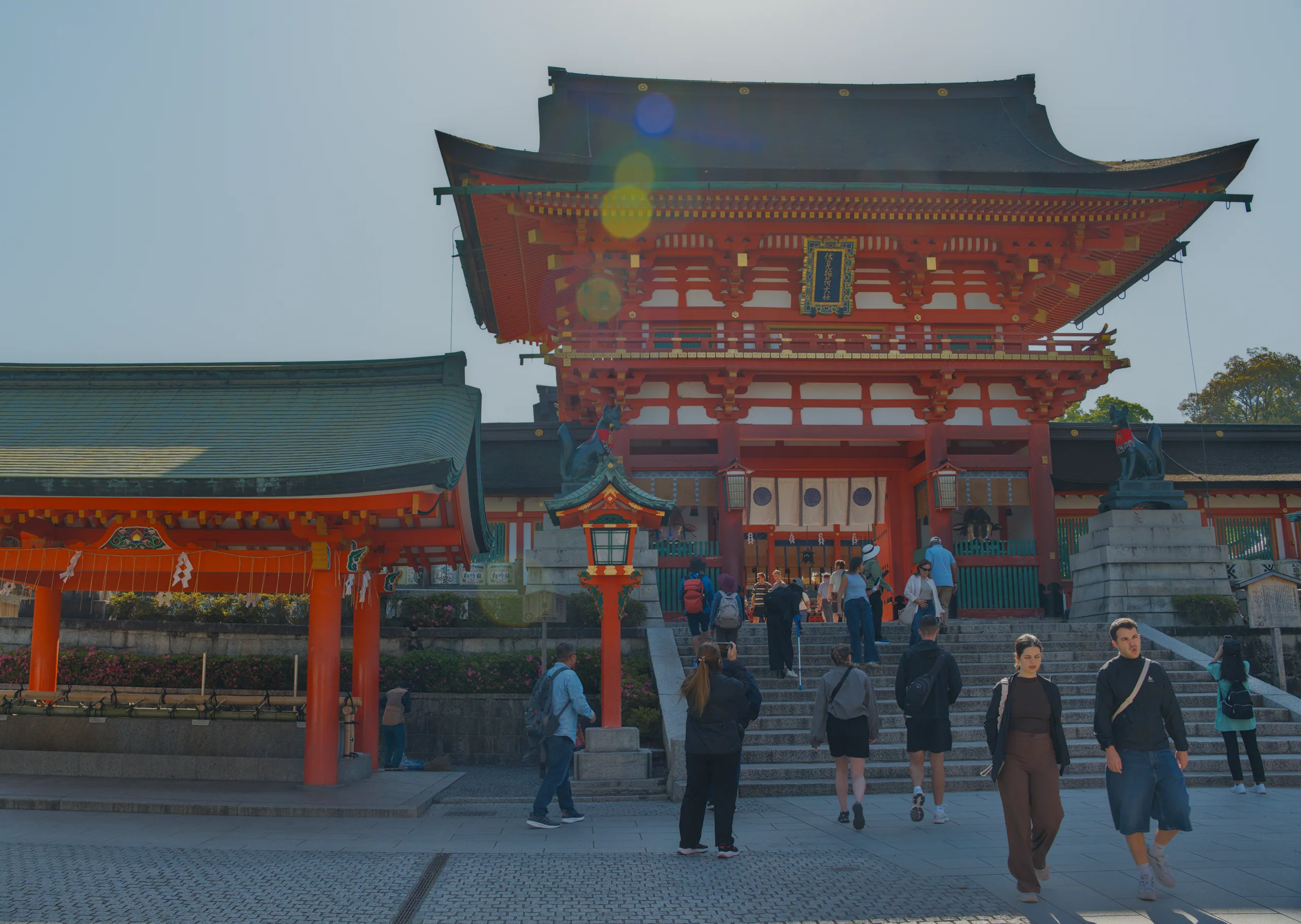 A red-roofed Japanese shrine building with curved eaves and people in the courtyard.