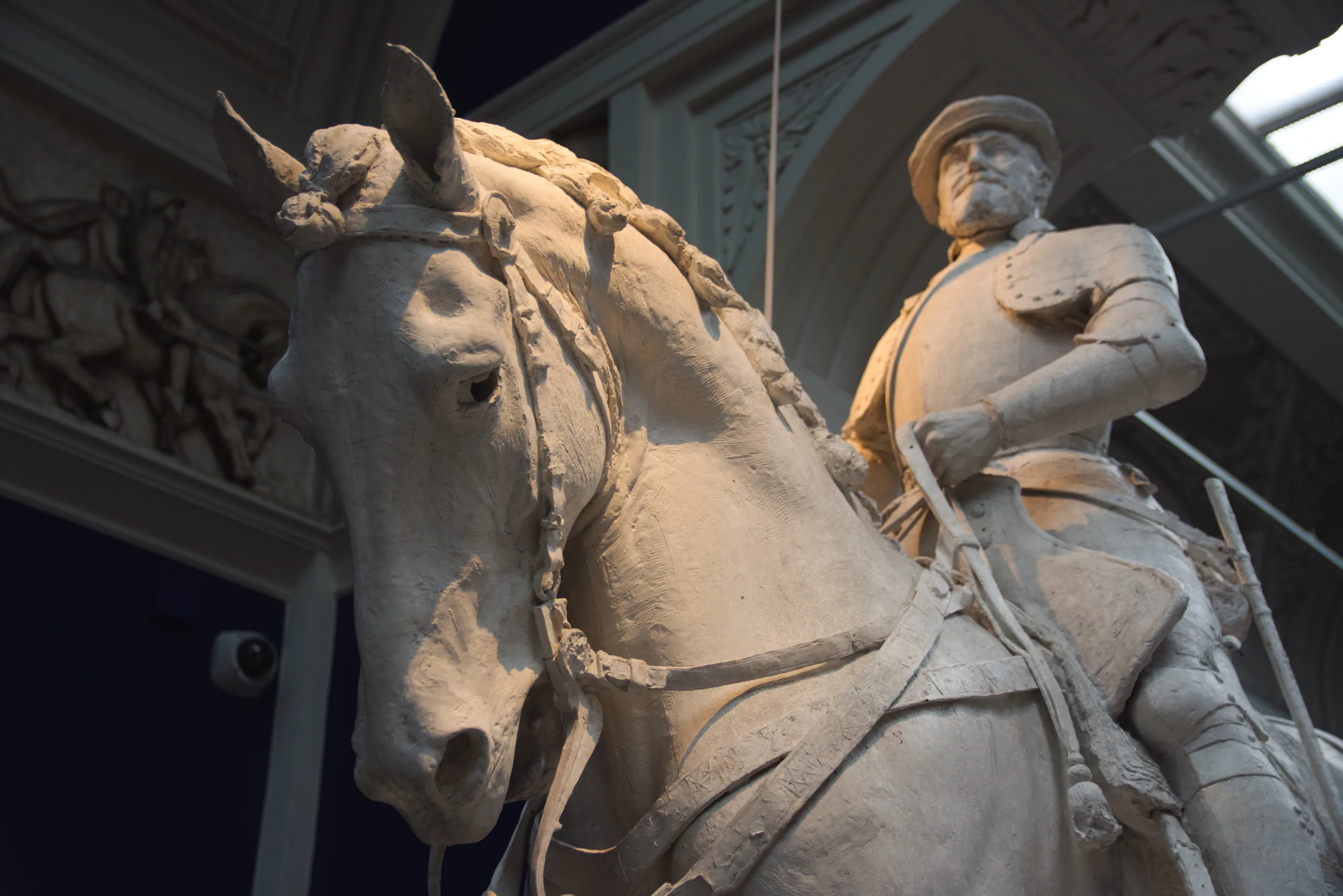 A plaster equestrian statue seen close from below, the horse's bridled head and the rider's arm against a vaulted gallery ceiling.
