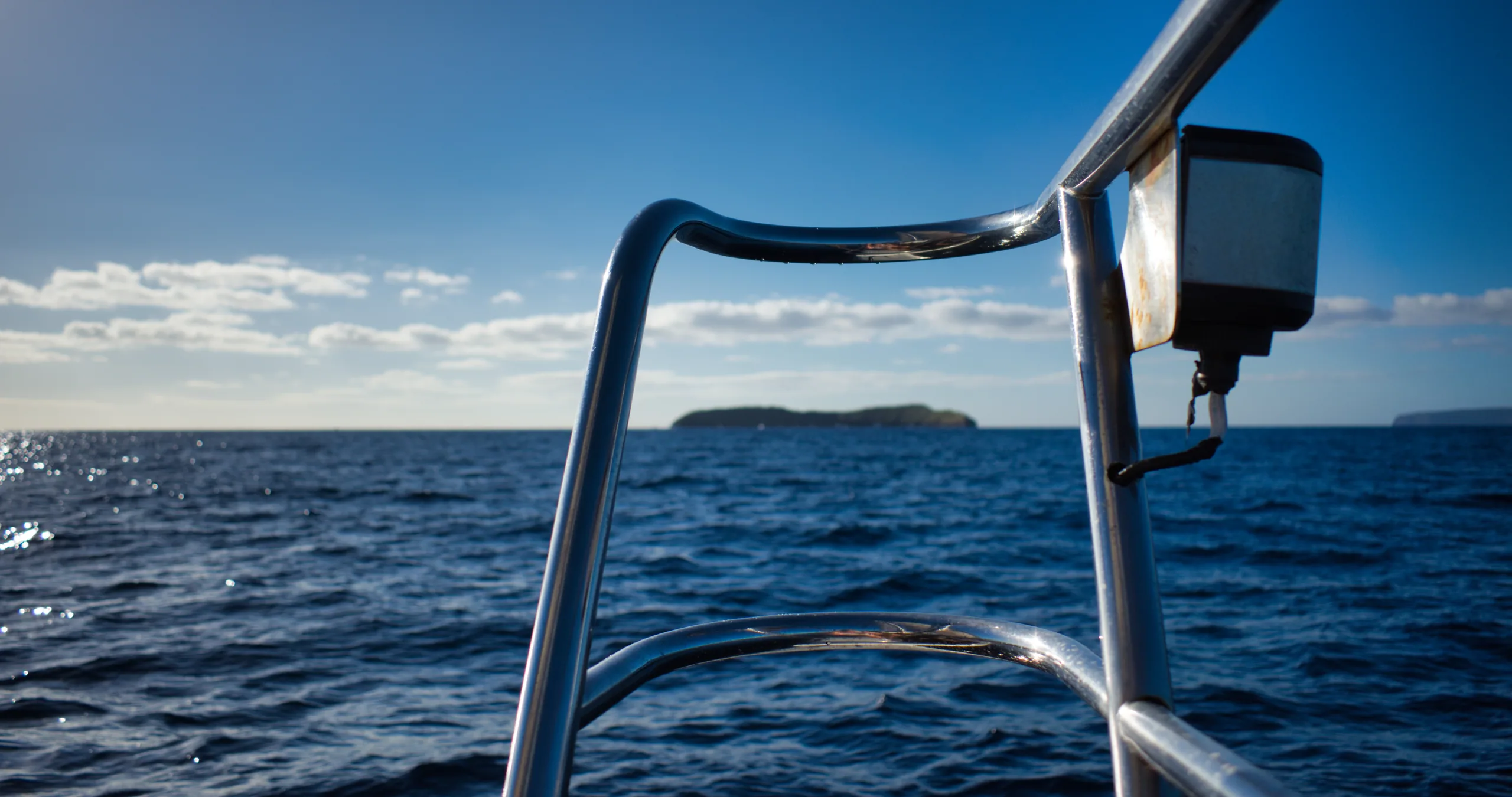 A stainless steel boat rail in the foreground with open ocean and a low island on the horizon beyond.