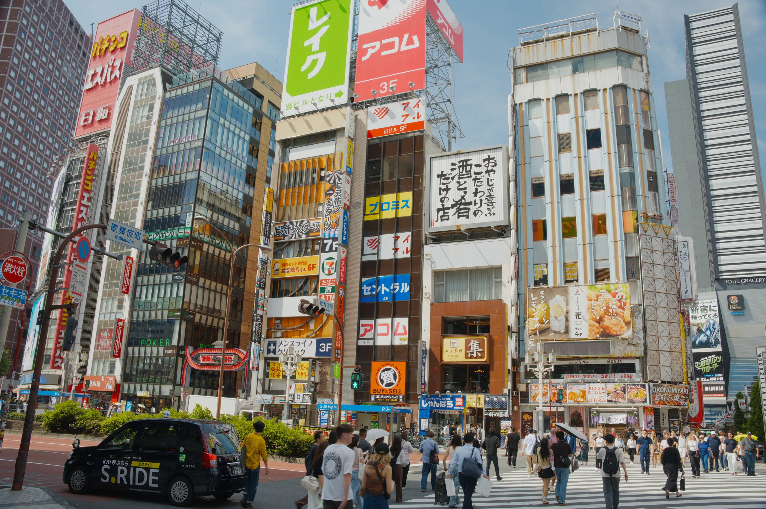 A dense commercial street with tall buildings covered in Japanese signage and pedestrians at a crosswalk.