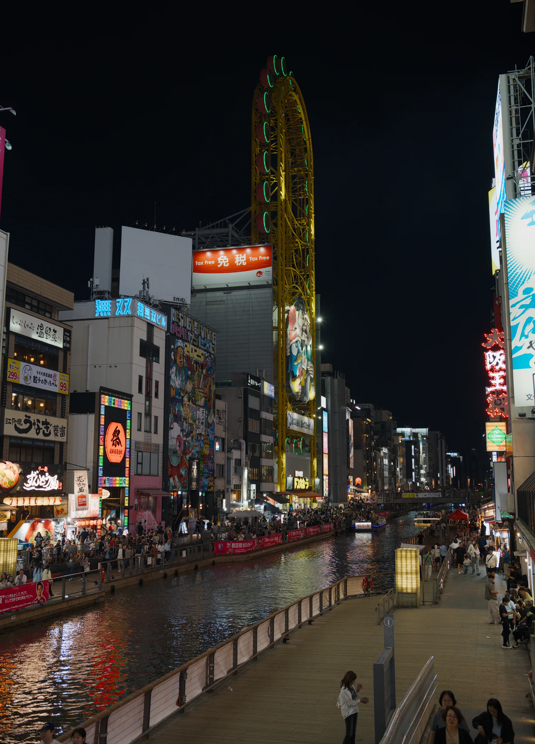 Neon-lit street beside a canal at night, with illuminated storefronts, signage, and pedestrians.
