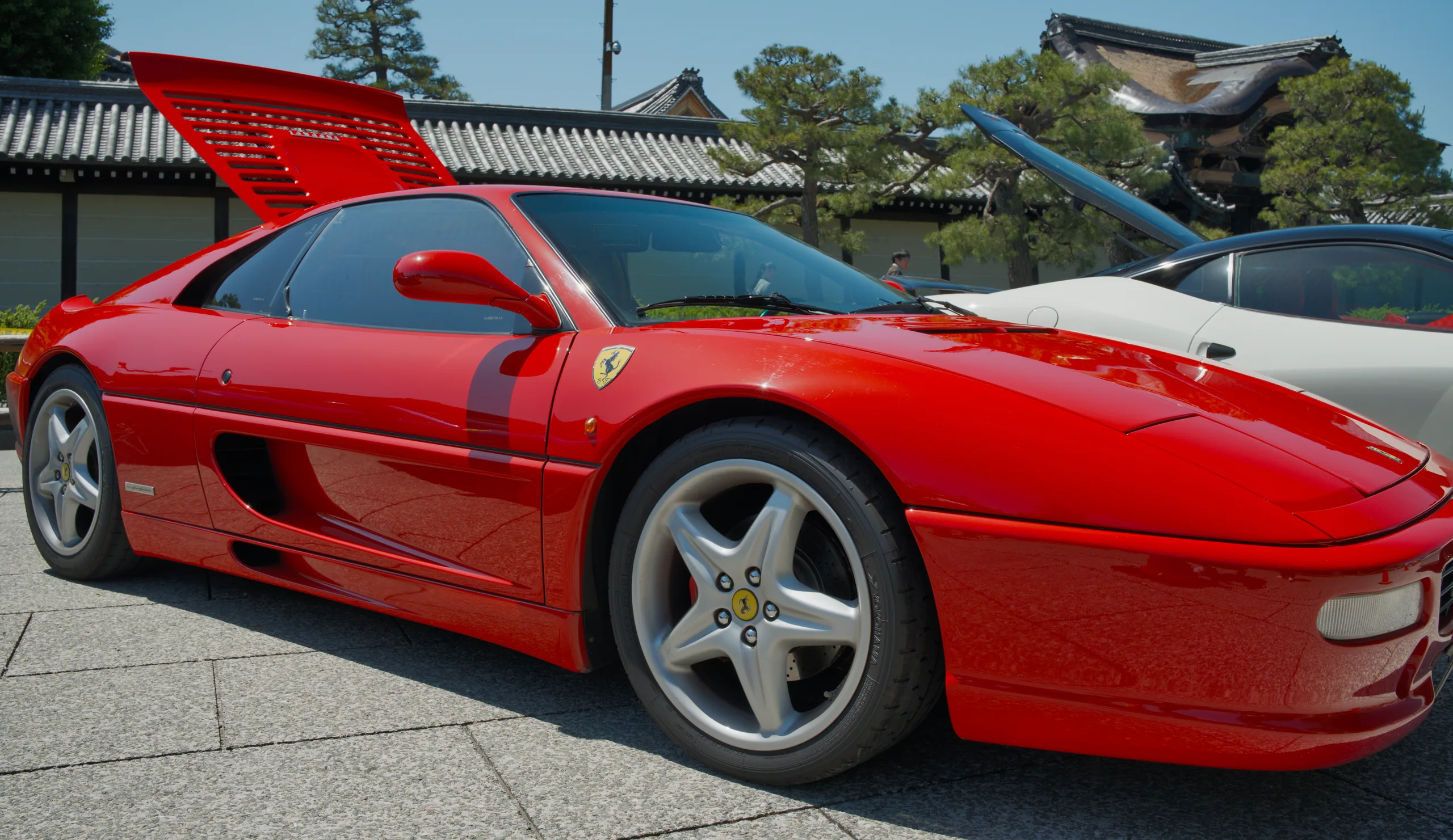A red Ferrari sports car parked next to a gray Porsche 911, with red-tiled roofed buildings in the background.