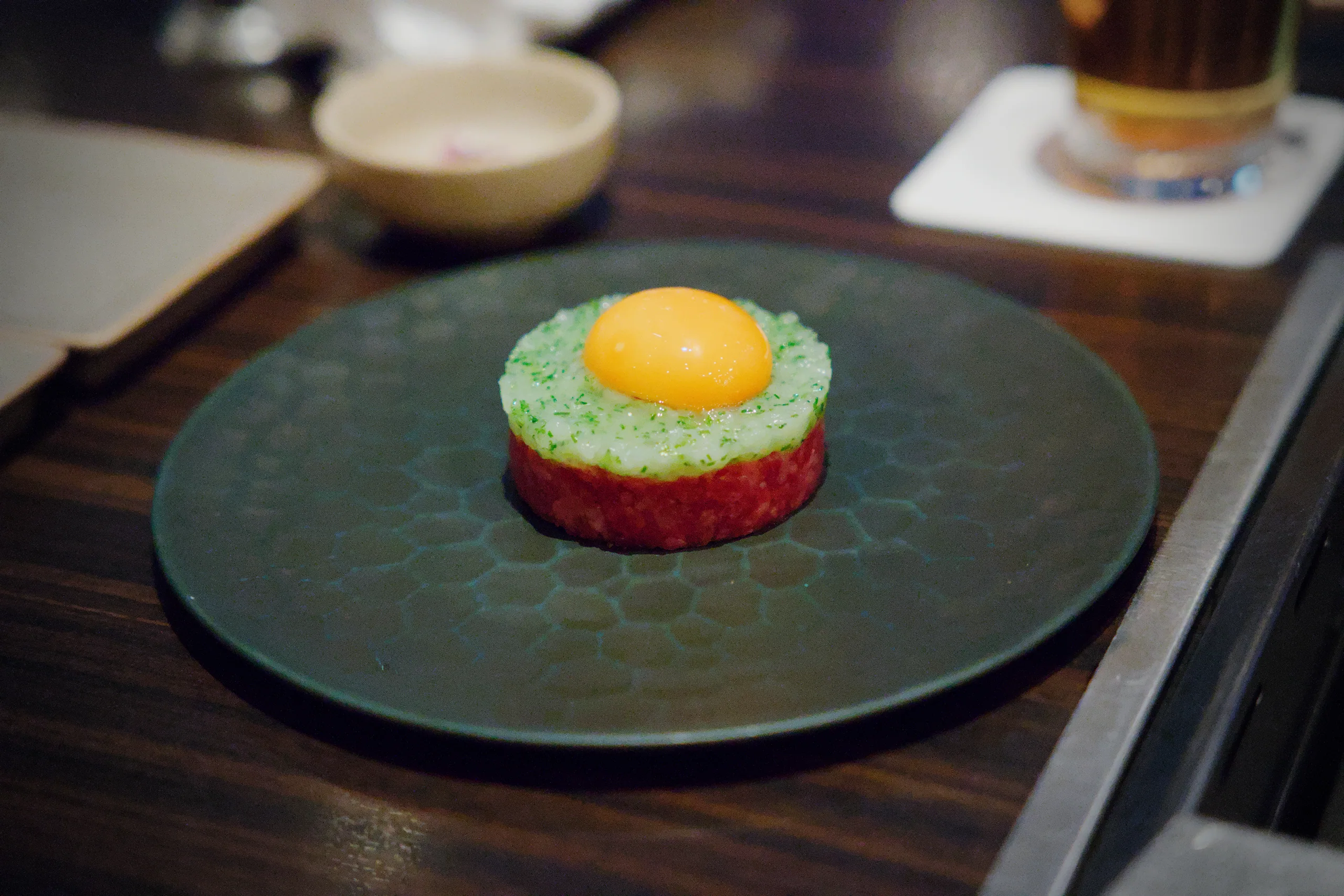 A bright raw egg yolk tops a plated composition on a matte green plate in a fine dining setting.