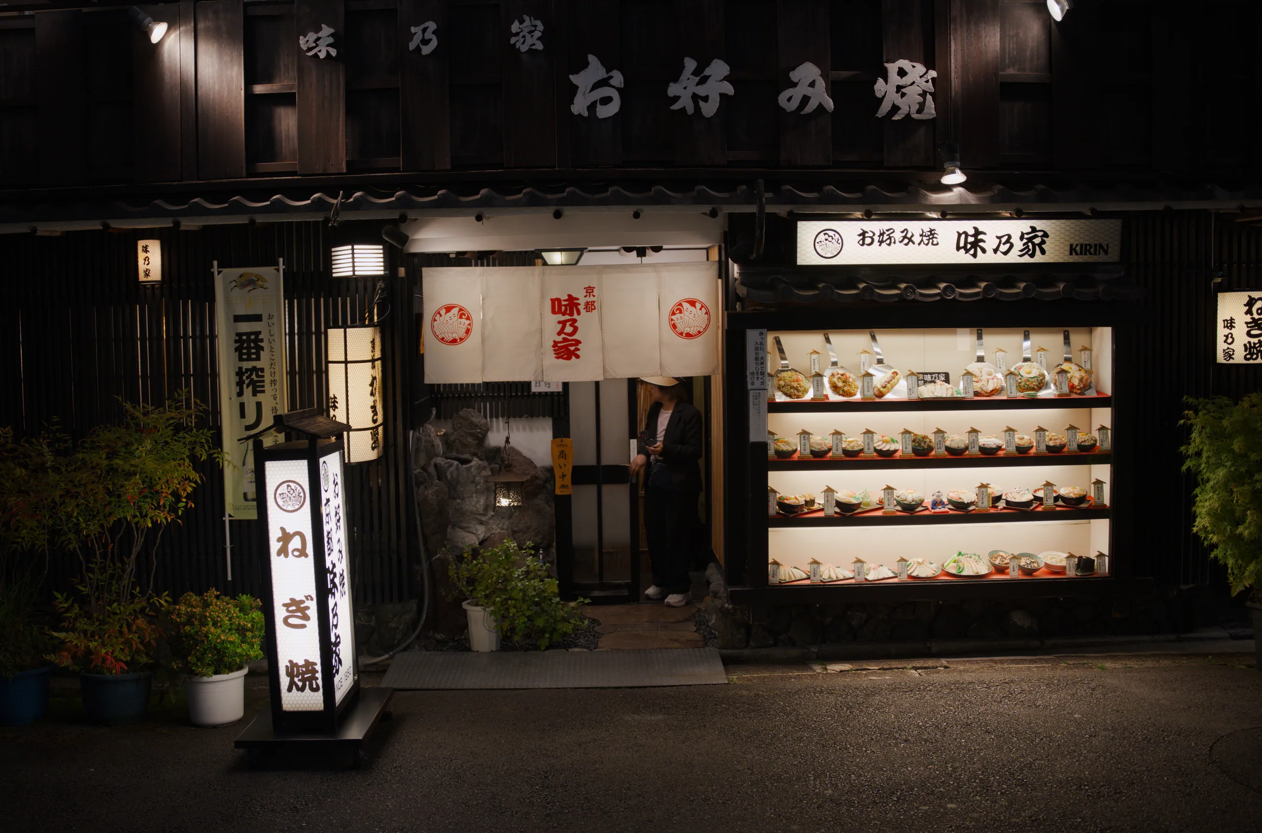 Japanese restaurant storefront at night with dark wood facade and lit window displaying grilled items.