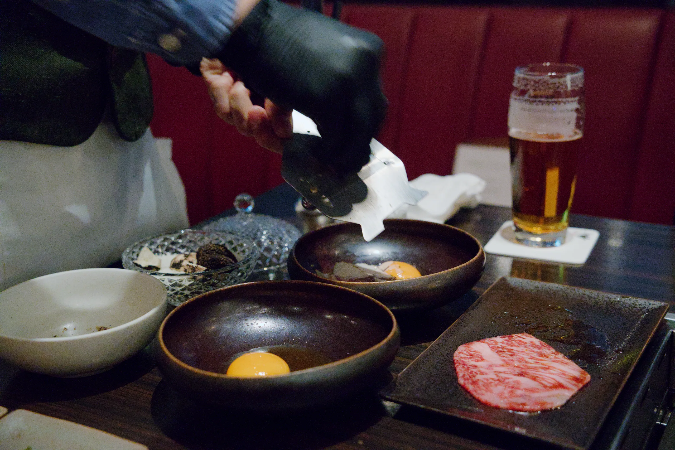 Dark bowls and slate plates holding eggs, sliced cured meat, and other components of a tasting menu are arranged on a table.