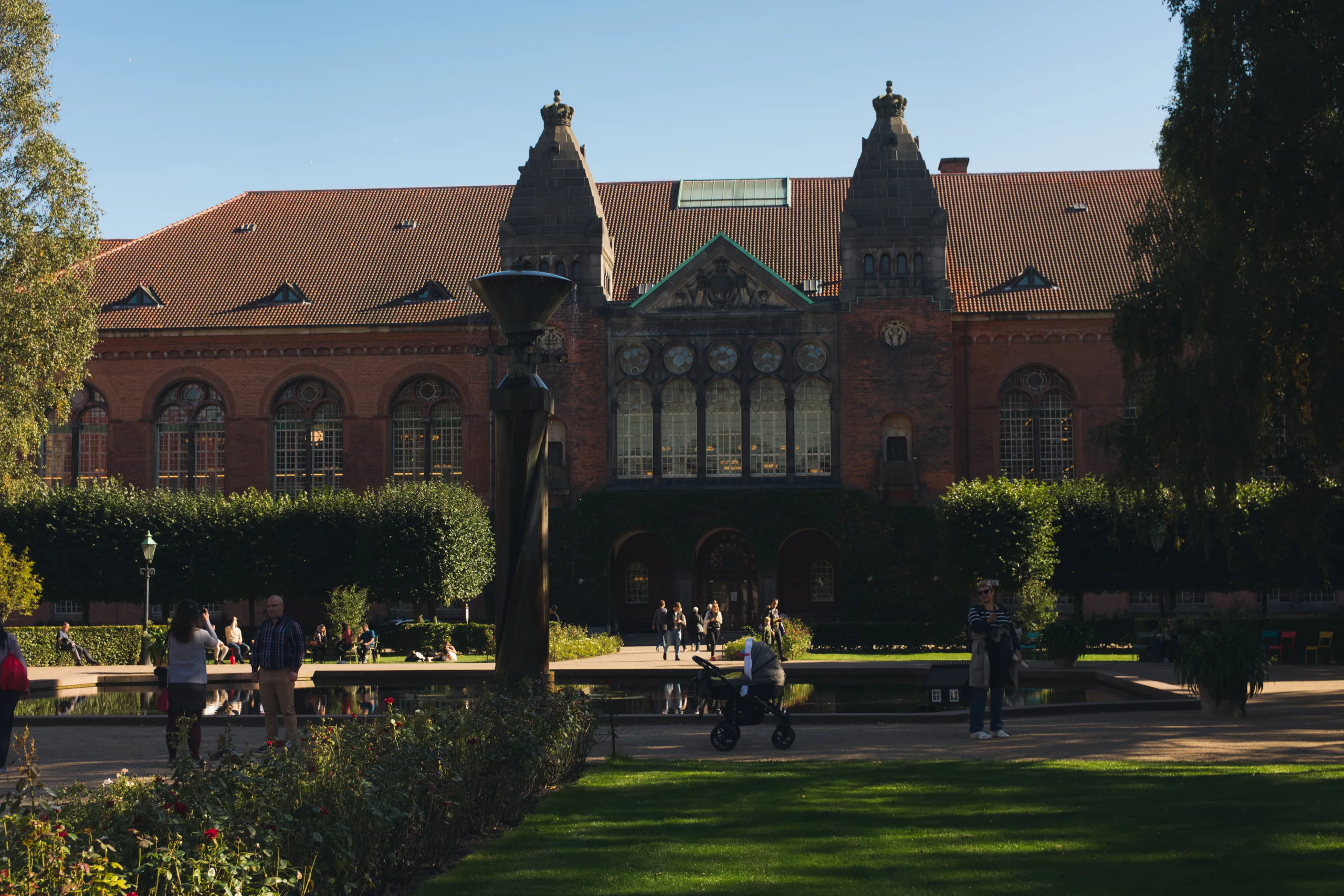 A red-brick museum building with twin towers and a decorated gable, seen across a sunlit public garden with a reflecting pool.