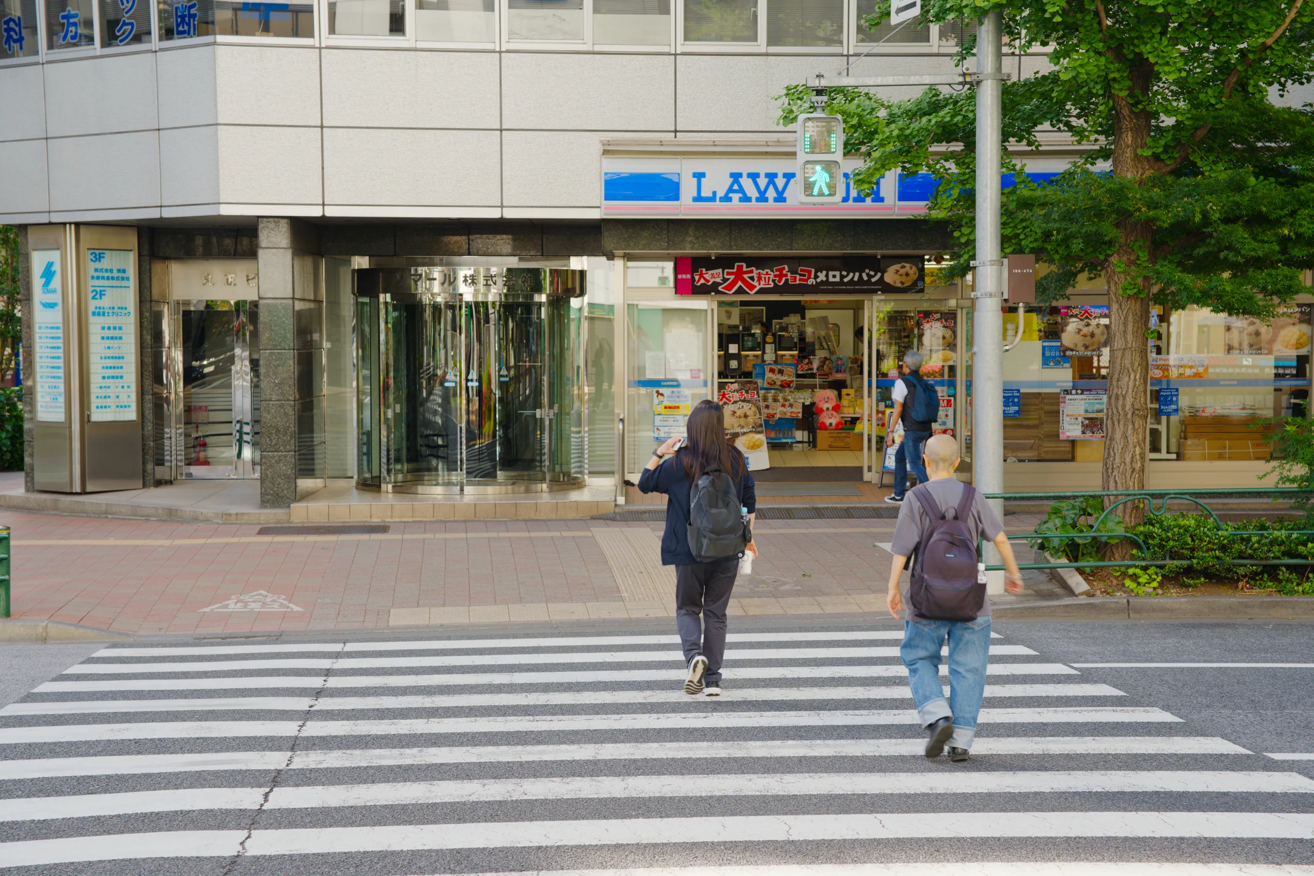 Pedestrians cross a zebra crossing in front of storefronts on a shopping street with trees.