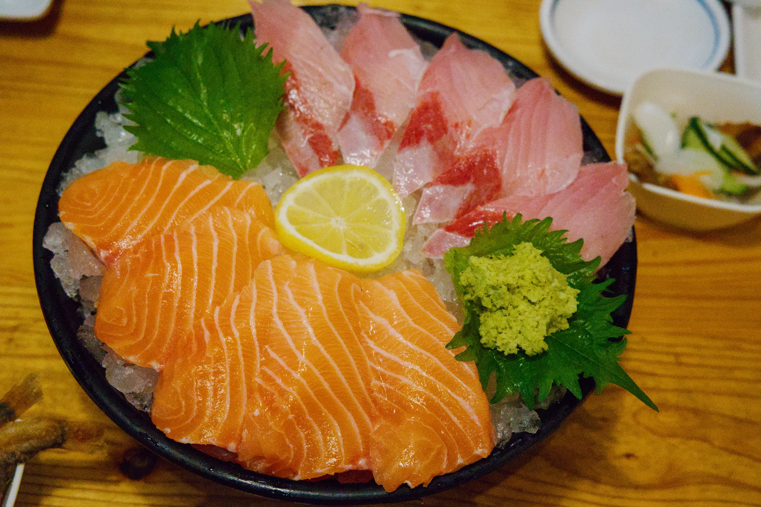 Sliced salmon and assorted sashimi arranged on a dark platter with shiso leaf garnish and lemon wedge.