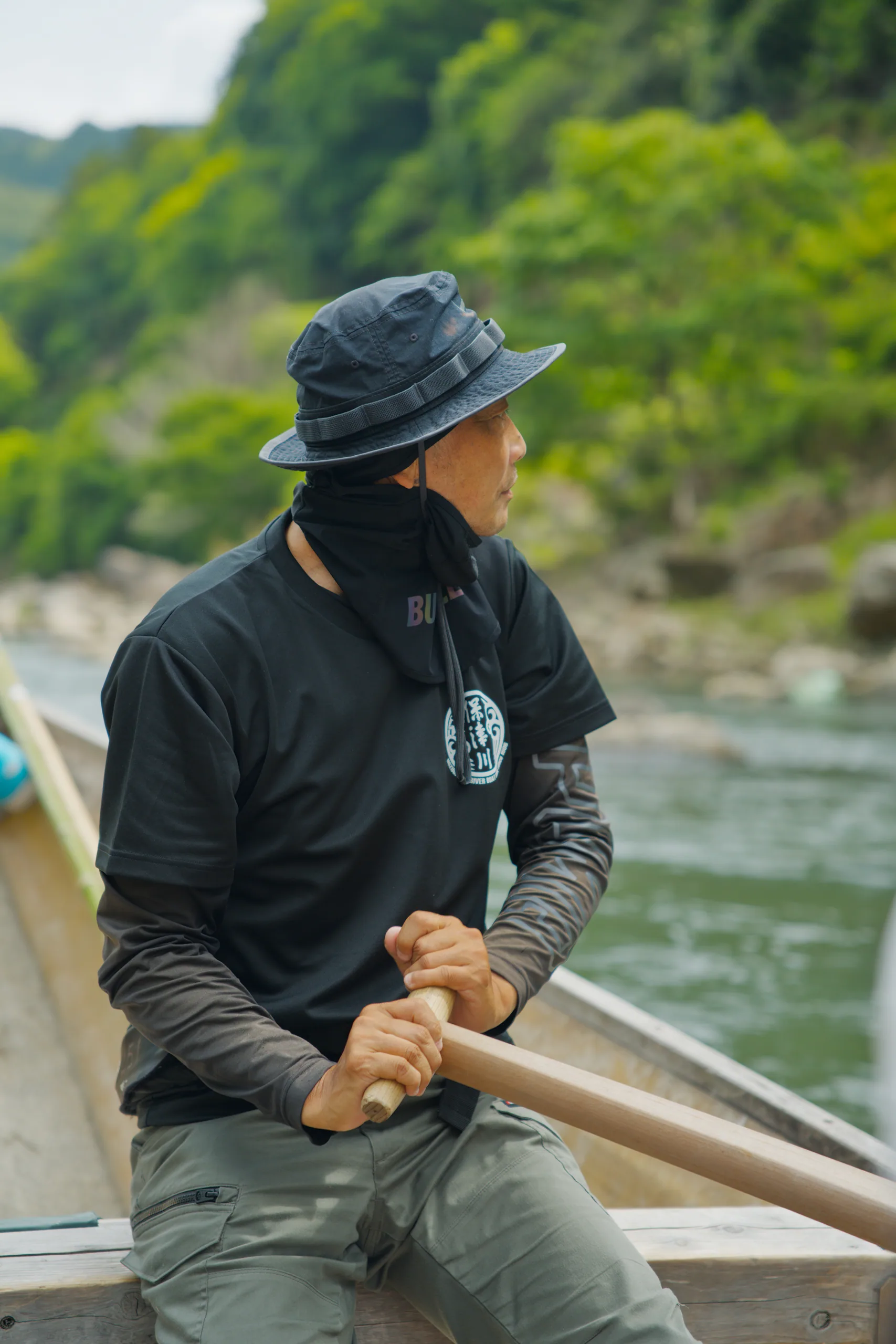 Person in a hat seated in a boat holding a paddle, steep forested hillsides visible across the water.